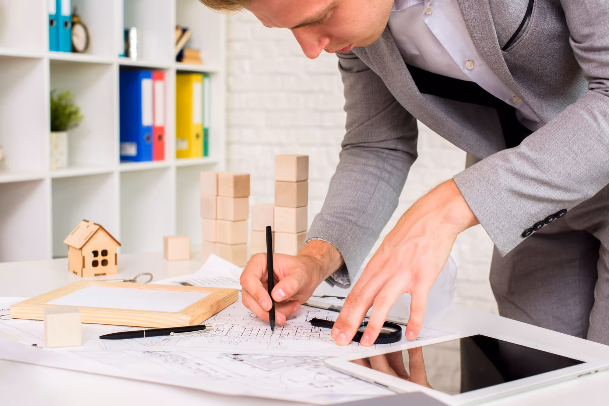 Close-up of a male architect or engineer in a grey suit leaning over a desk, drawing or marking a blueprint with a black pen, with a magnifying glass, a tablet, a small wooden house model, and wooden blocks visible on the table.