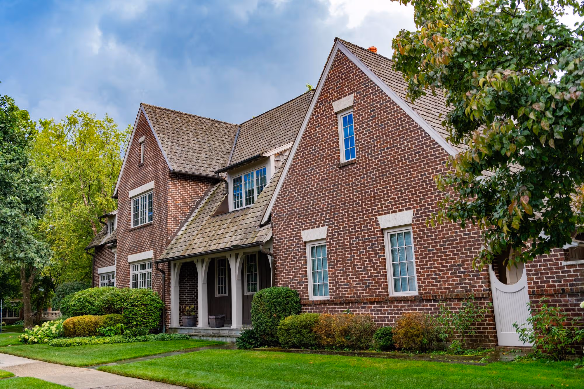 Exterior of a traditional, two-story house with a steep gable roof, dark red brick facade, white window trim, and a small covered front porch, surrounded by a manicured green lawn and mature trees.
