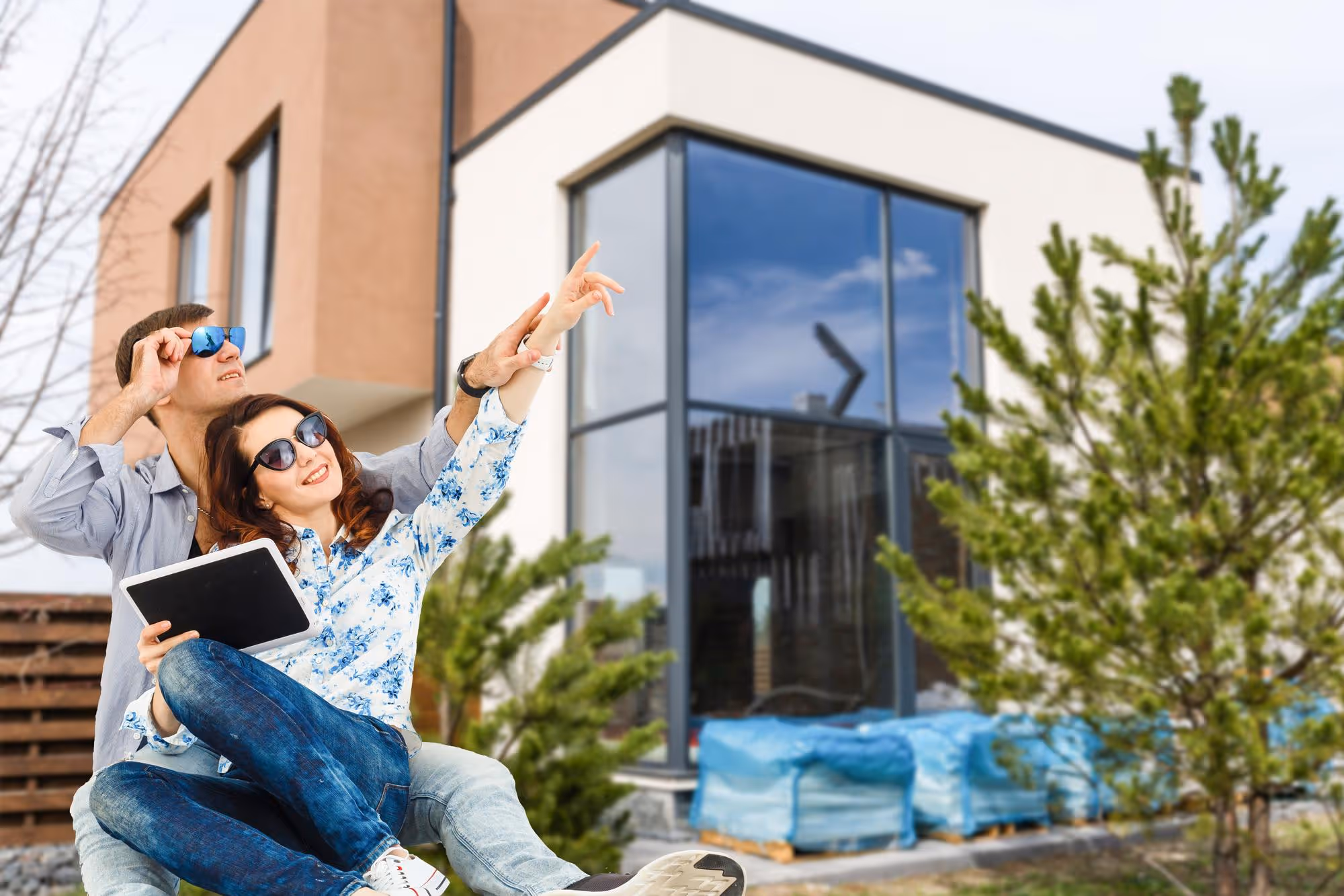 A smiling young couple wearing sunglasses and casual clothes sit together outside a modern, newly constructed home, looking up and pointing at the house while holding a digital tablet. Construction materials covered in blue tarp are visible in the yard.