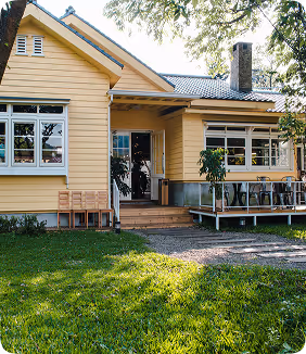 Yellow wooden house with large windows, front porch, and green lawn surrounded by trees, creating a cozy and inviting residential exterior.