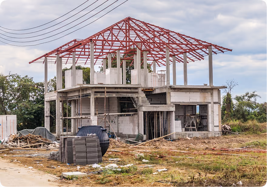 Exterior view of a two-story house under construction in a rural setting, with the concrete walls and pillars partially built, and the red metal truss framework for the roof fully installed. Construction materials are scattered in the foreground.