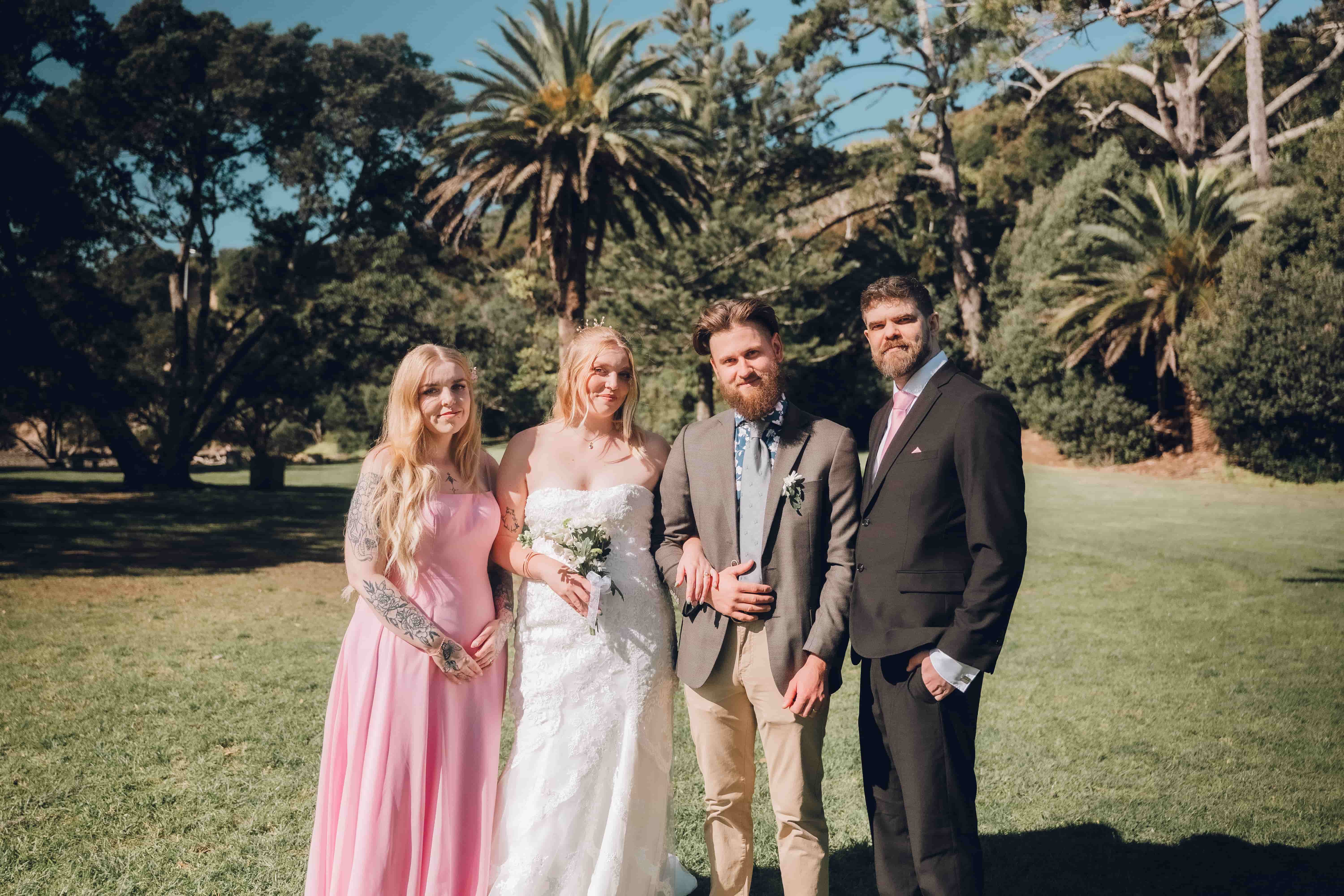 Bride and groom standing with their bridal party under palm trees at a sunny Auckland wedding, photographed by Vantirra