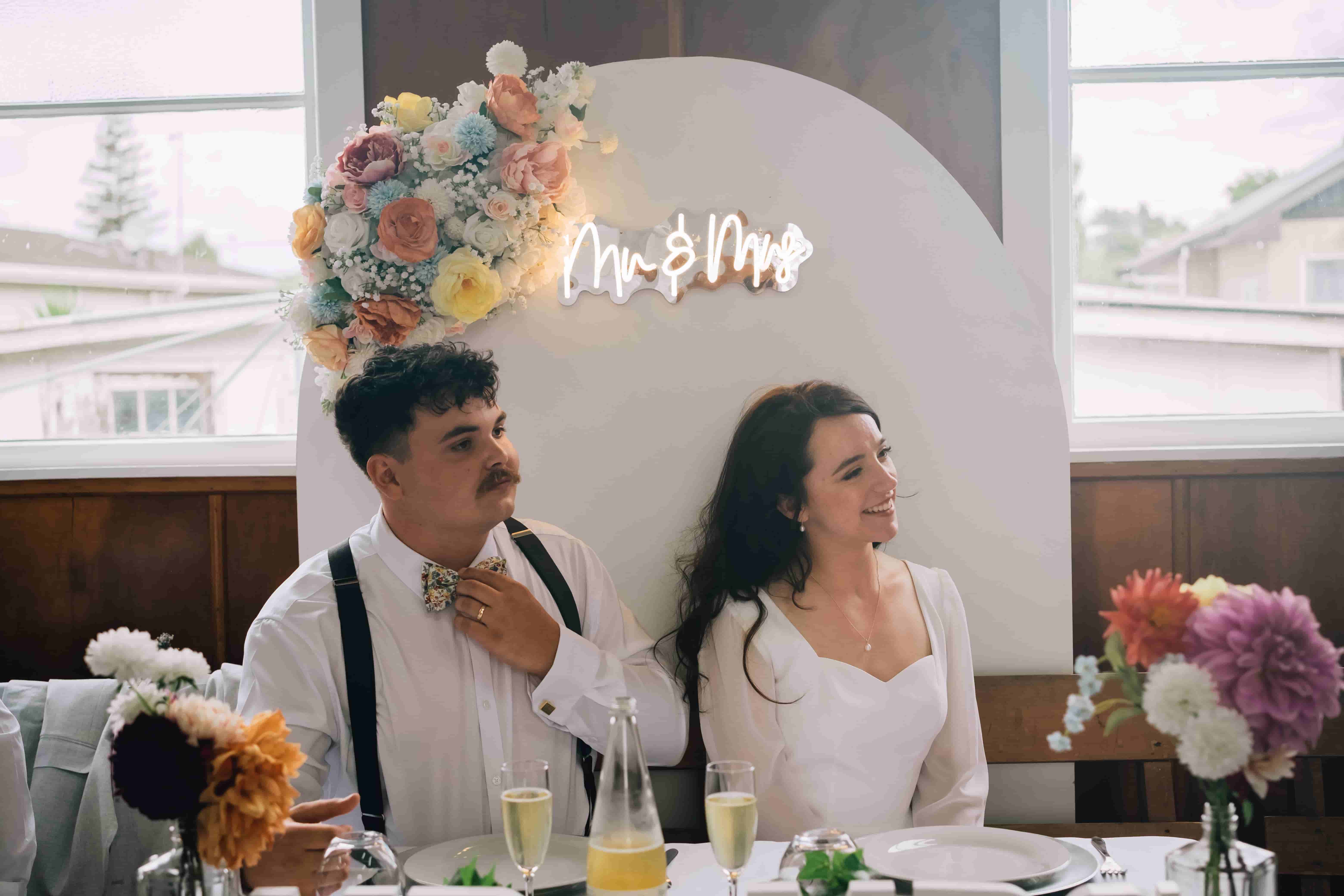 A bride and groom seated at their reception table in front of a floral arch and a neon Mr & Mrs sign, photographed by Vantirra