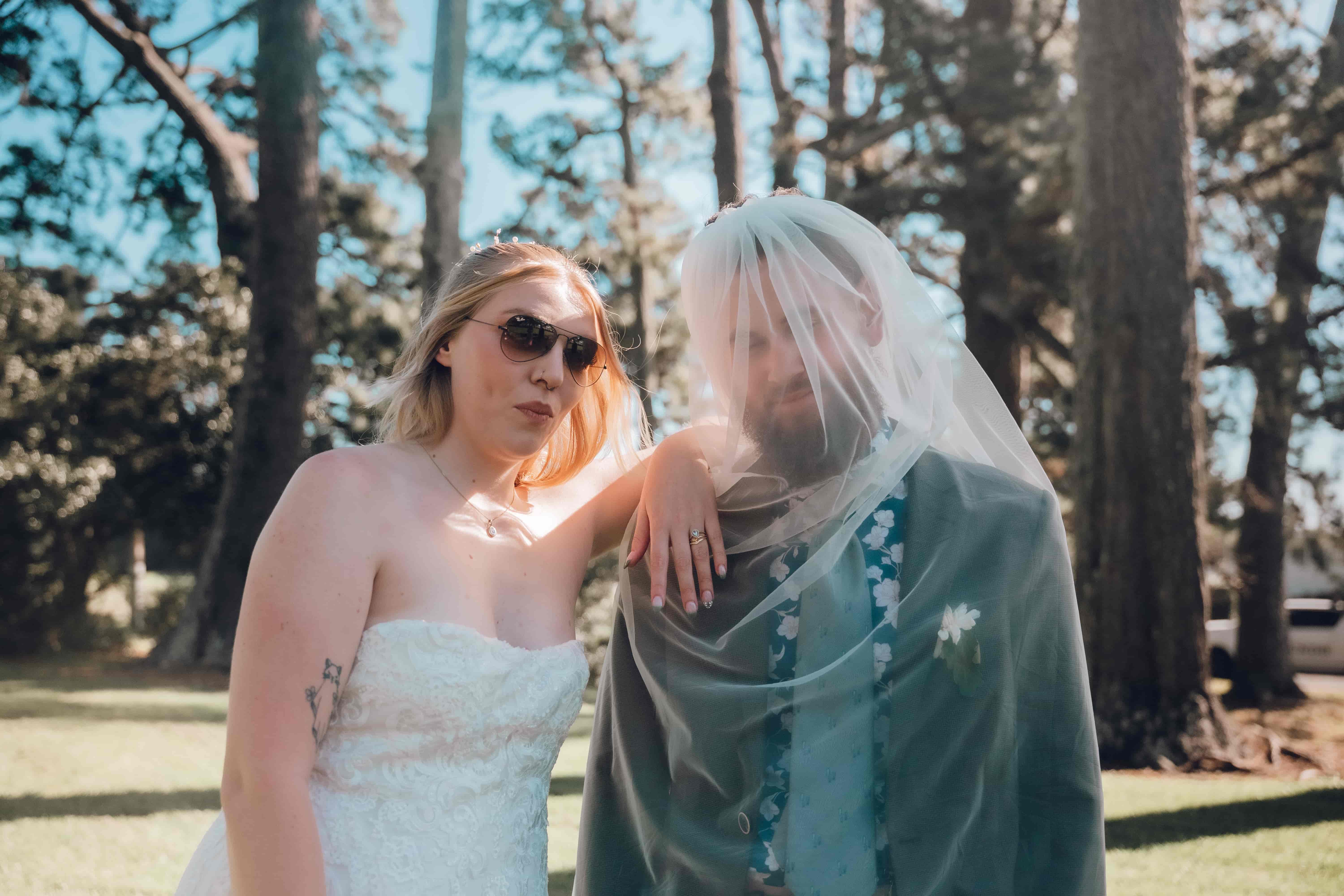 A bride in sunglasses resting her arm on the groom, who has her veil draped over his head, laughing together under pine trees, photographed by Vantirra