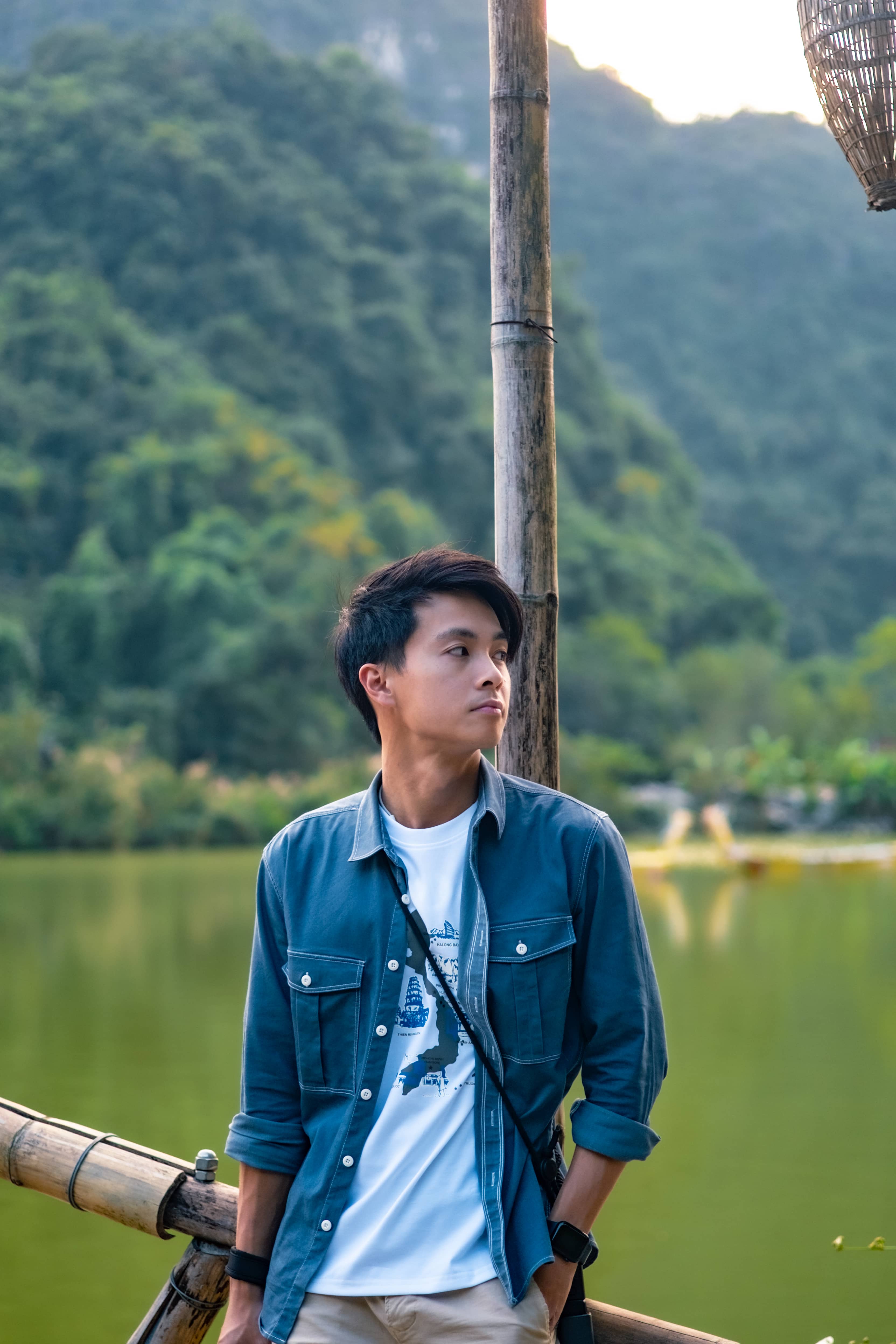 Close portrait of a man in a denim jacket looking aside by a lake, a woven lantern and green hills behind