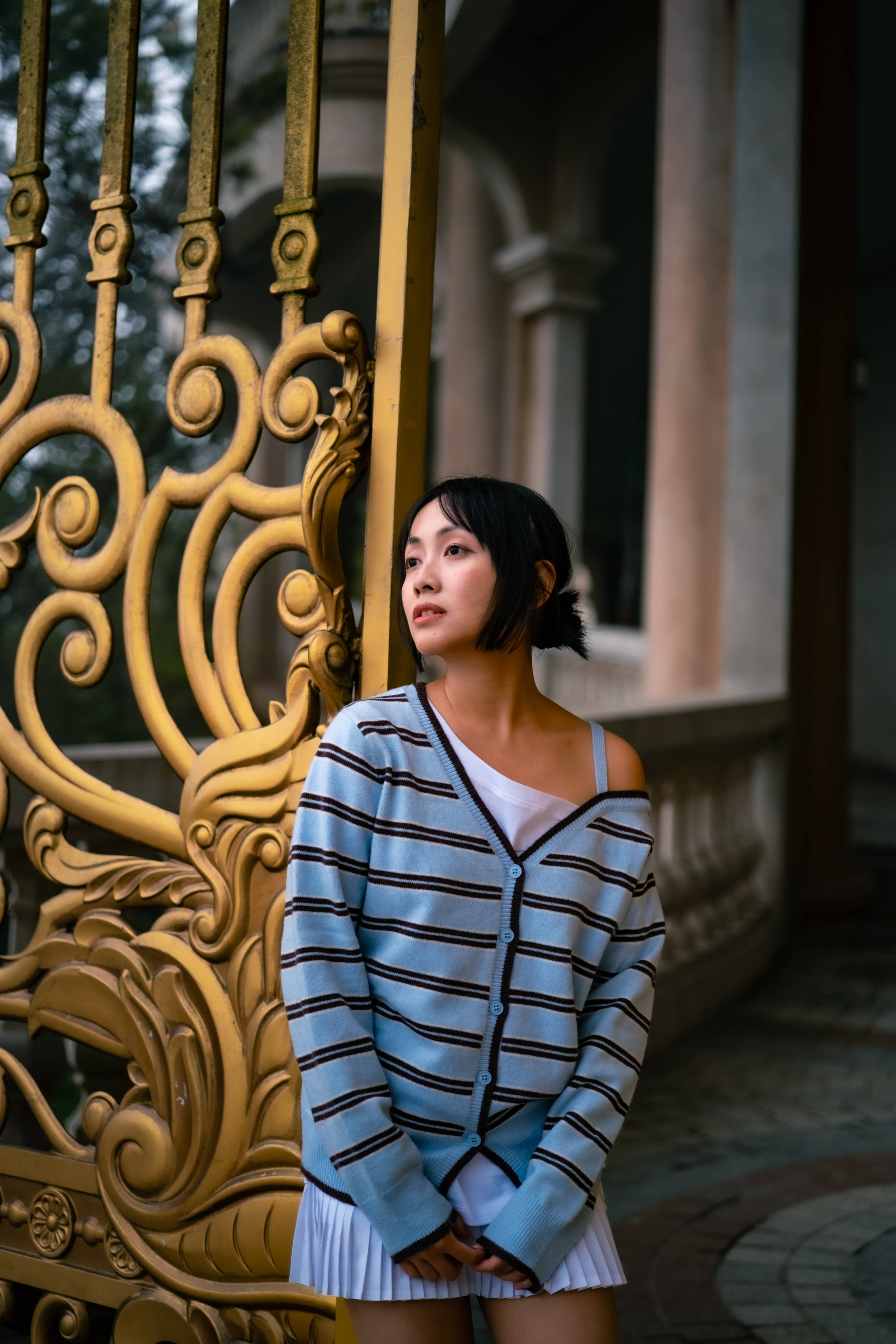 Woman in a blue striped cardigan and white pleated skirt standing by an ornate gold gate at a colonnaded building, Vietnam