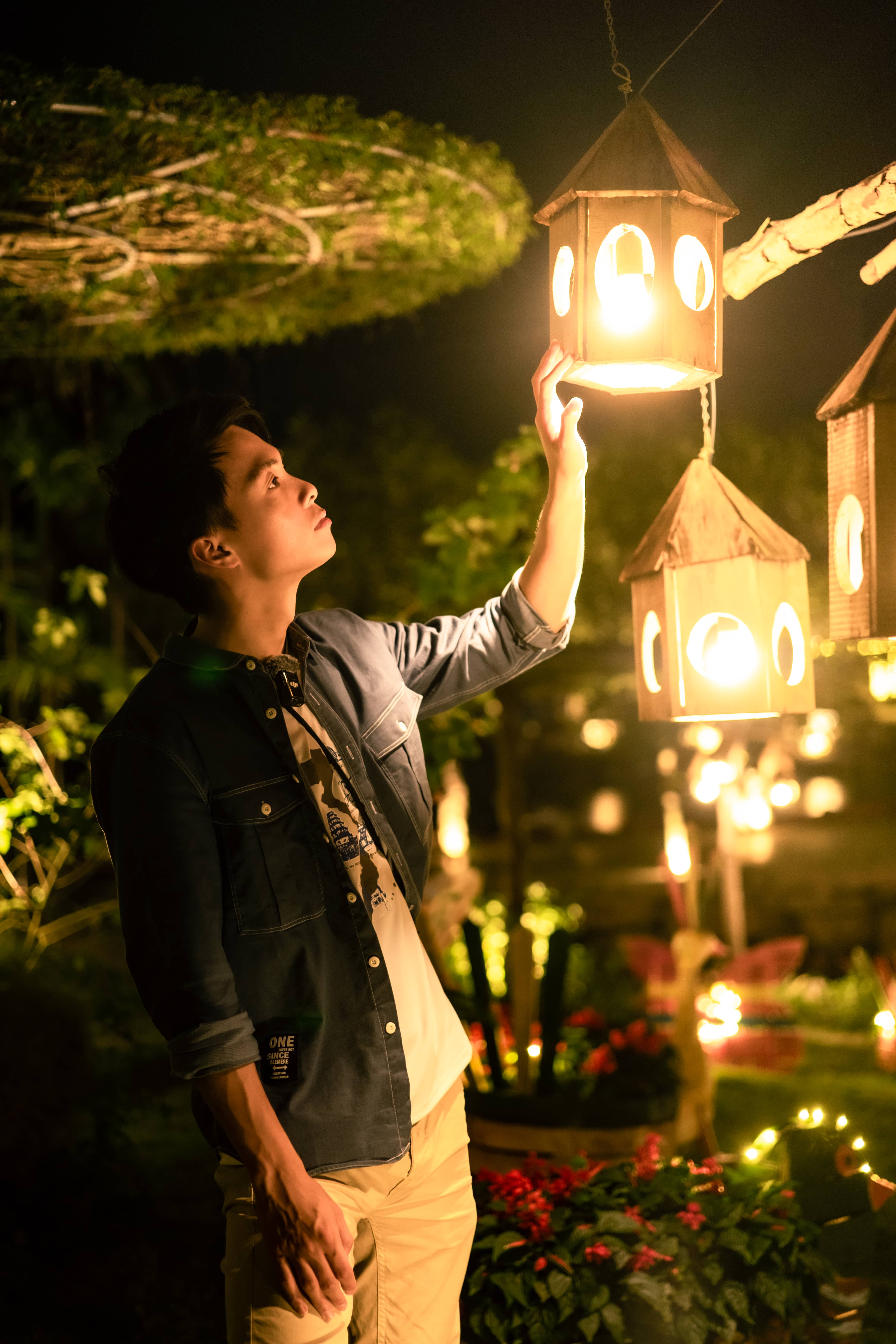 Man in a denim jacket reaching up to a glowing wooden lantern house hanging in a garden at night