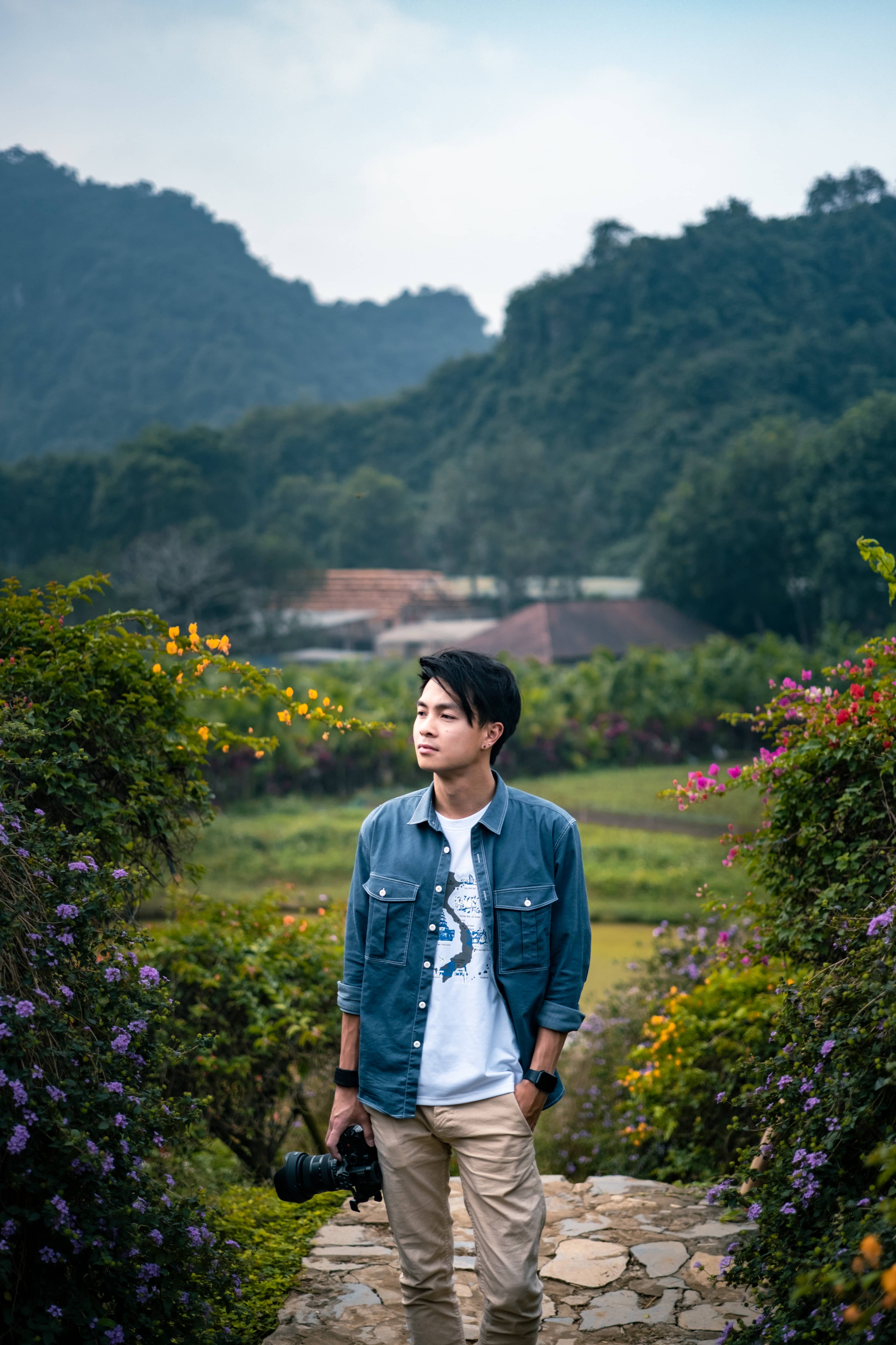 Man holding a camera on a stone path between flowering shrubs, red roofs and green peaks behind