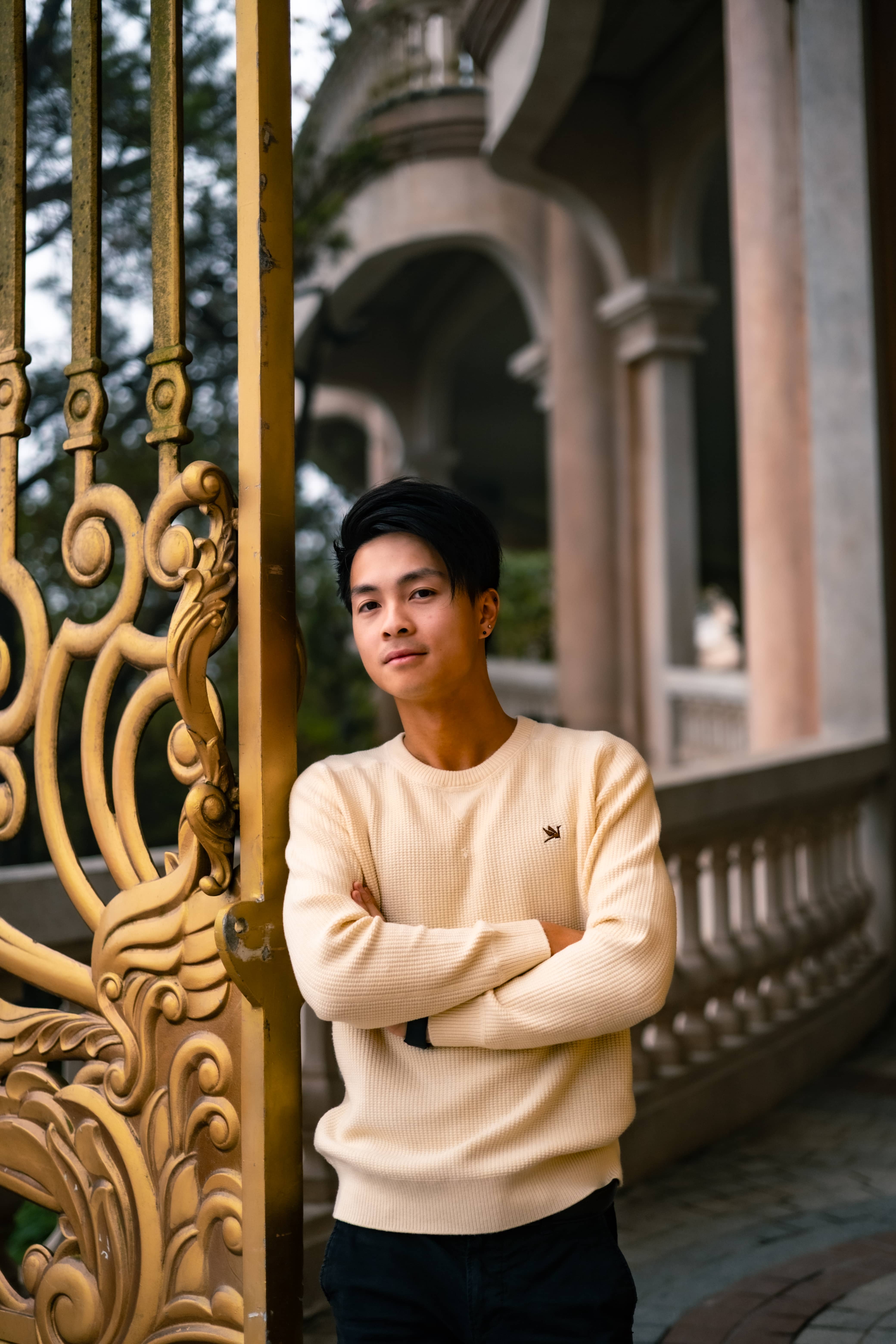 Young man in a cream knit sweater, arms crossed, leaning by an ornate gold gate at a colonnaded building, Vietnam