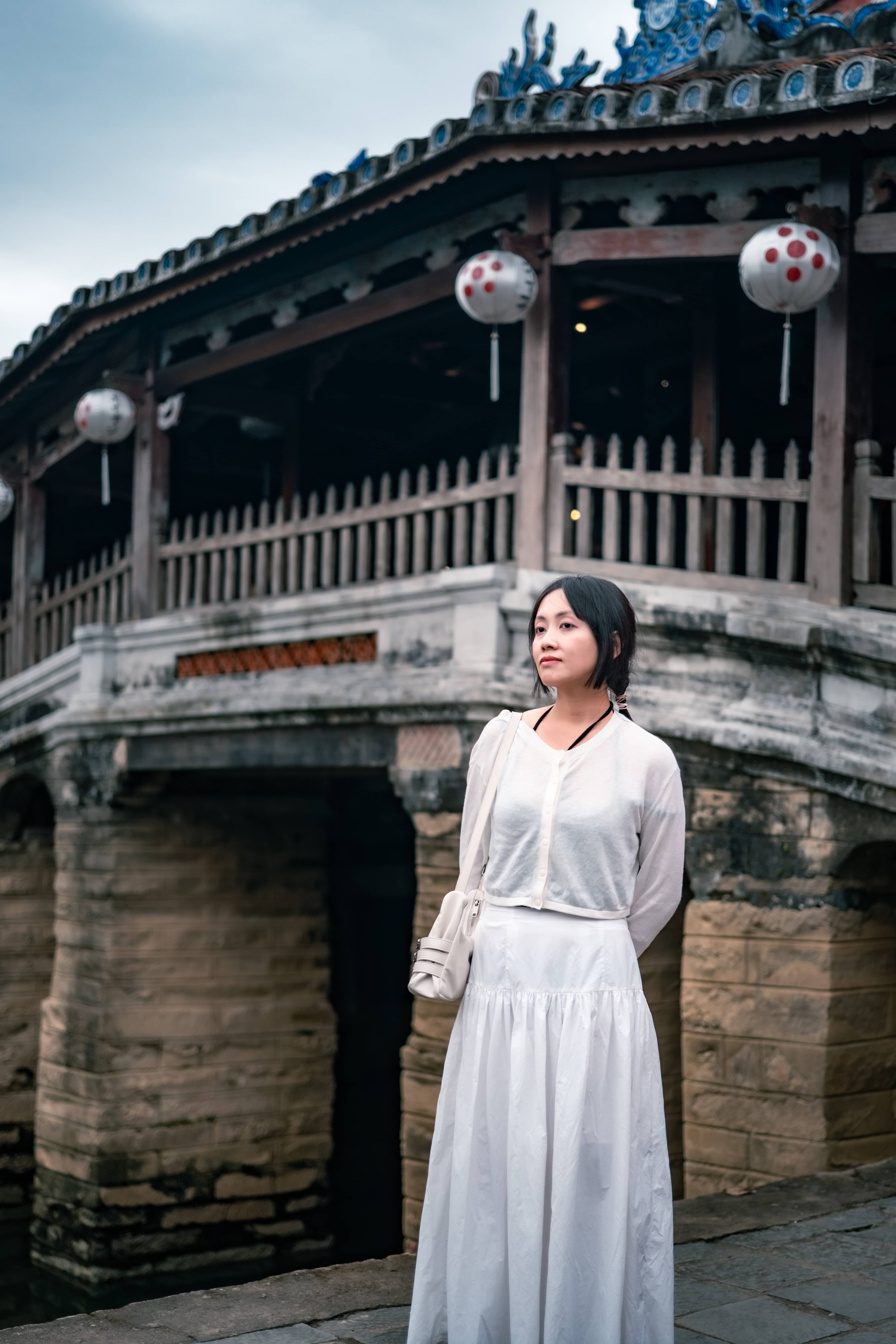 Woman in a long white dress standing before an old covered bridge with a tiled roof and paper lanterns