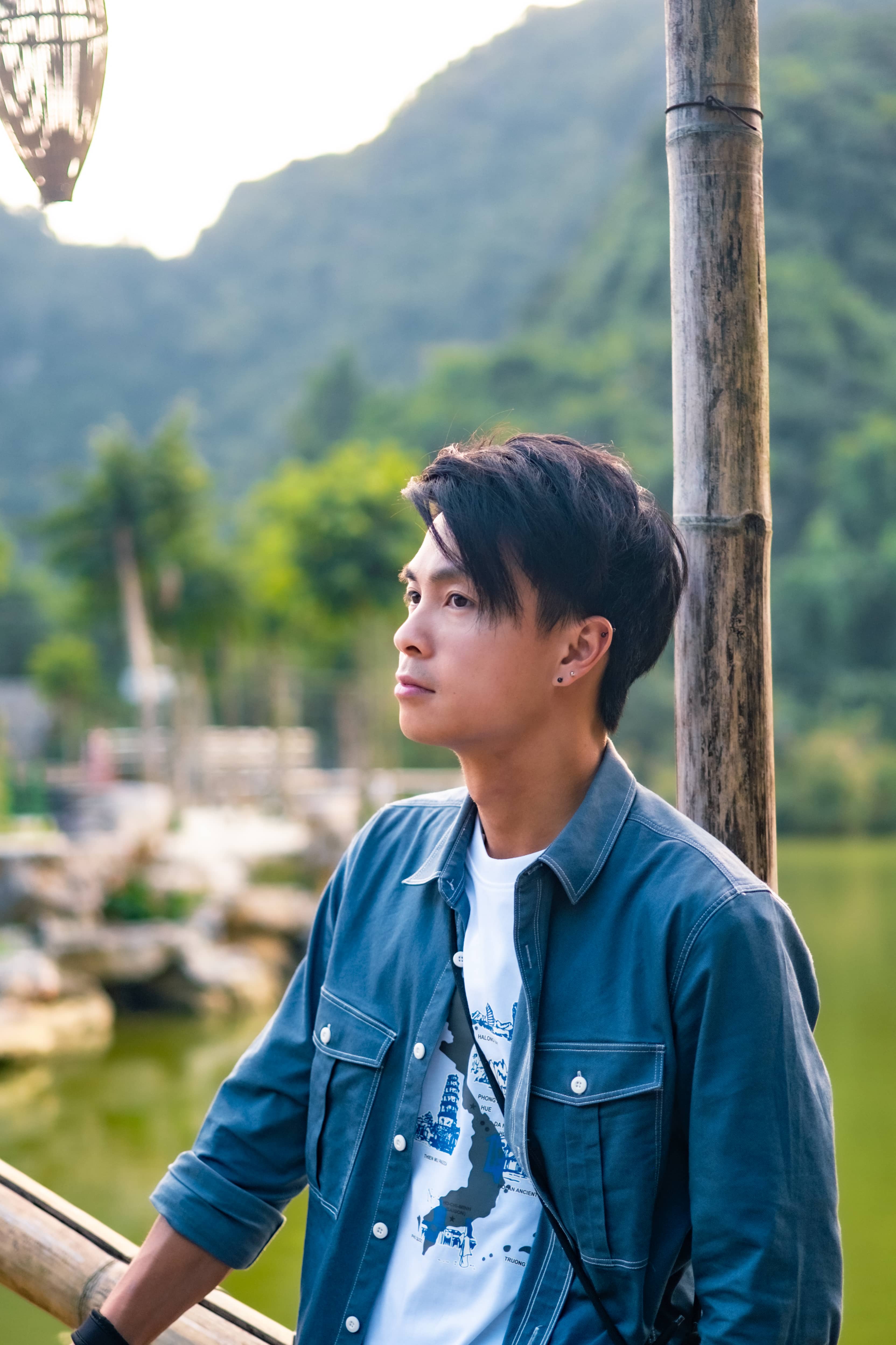 Man in a denim jacket leaning on a bamboo pole, looking out over a lake beneath misty green mountains