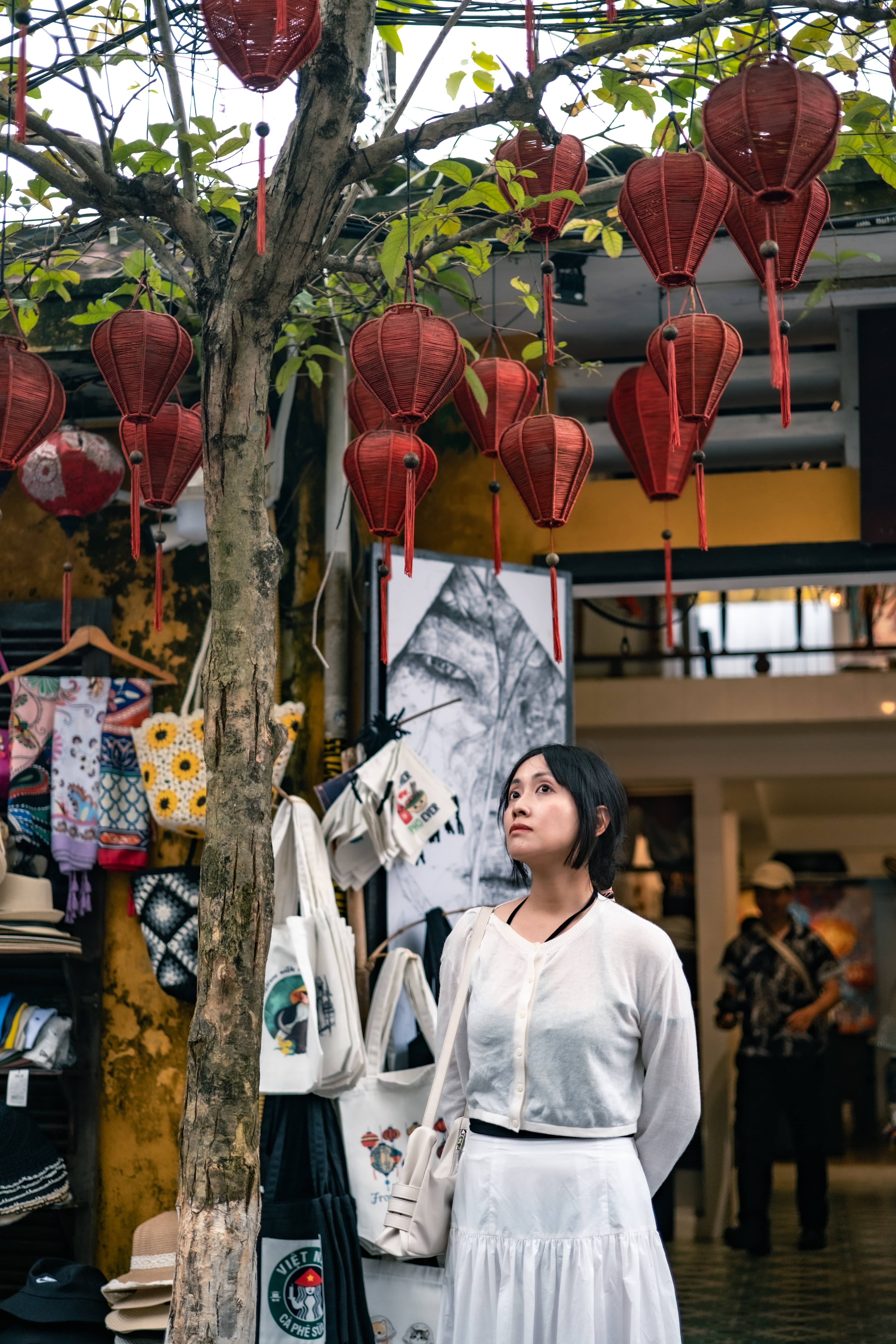 Woman in a white blouse and skirt beside a shop hung with red silk lanterns and souvenir bags, Vietnam