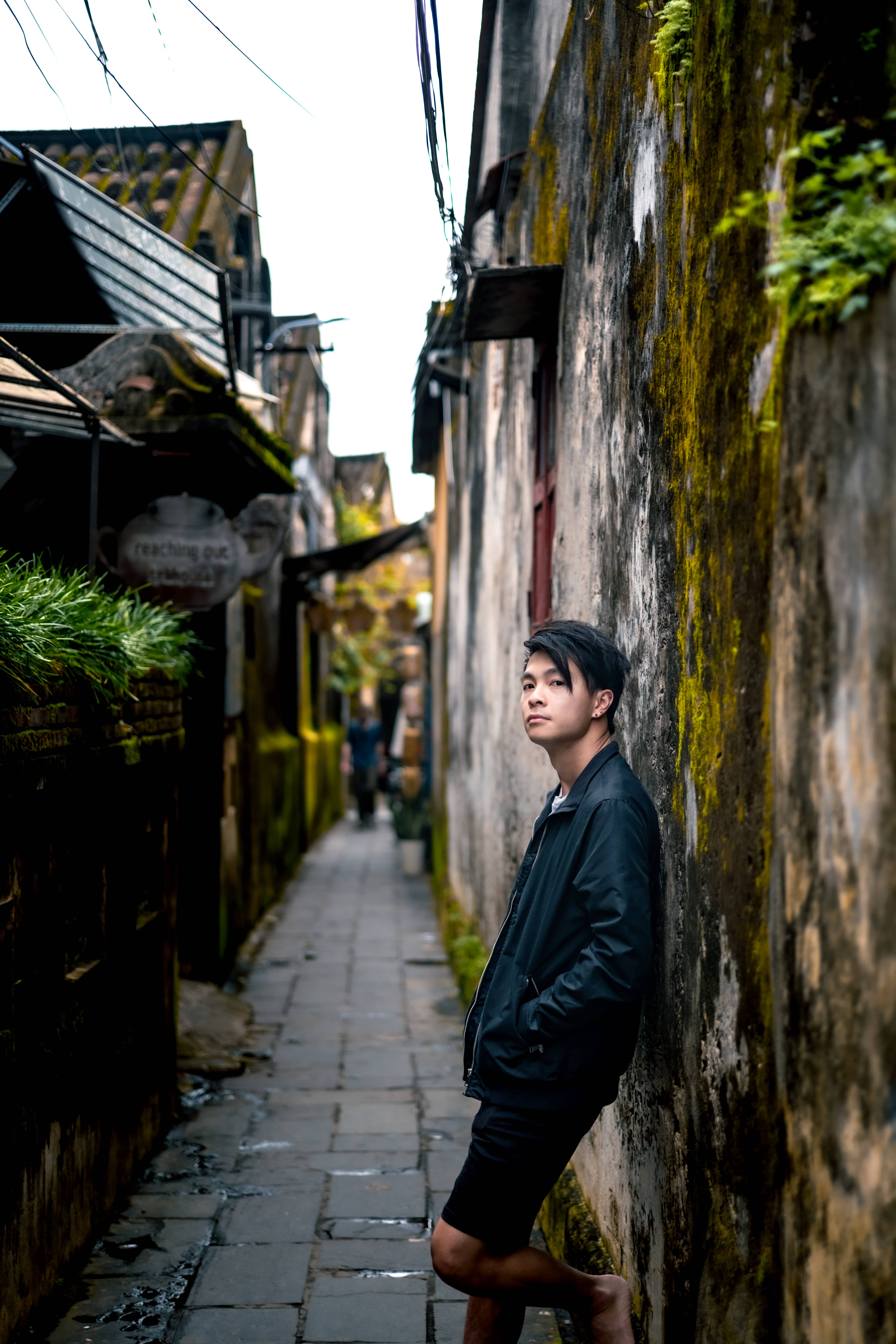 Man in a black jacket leaning on a mossy weathered wall in a narrow paved alley, Vietnam