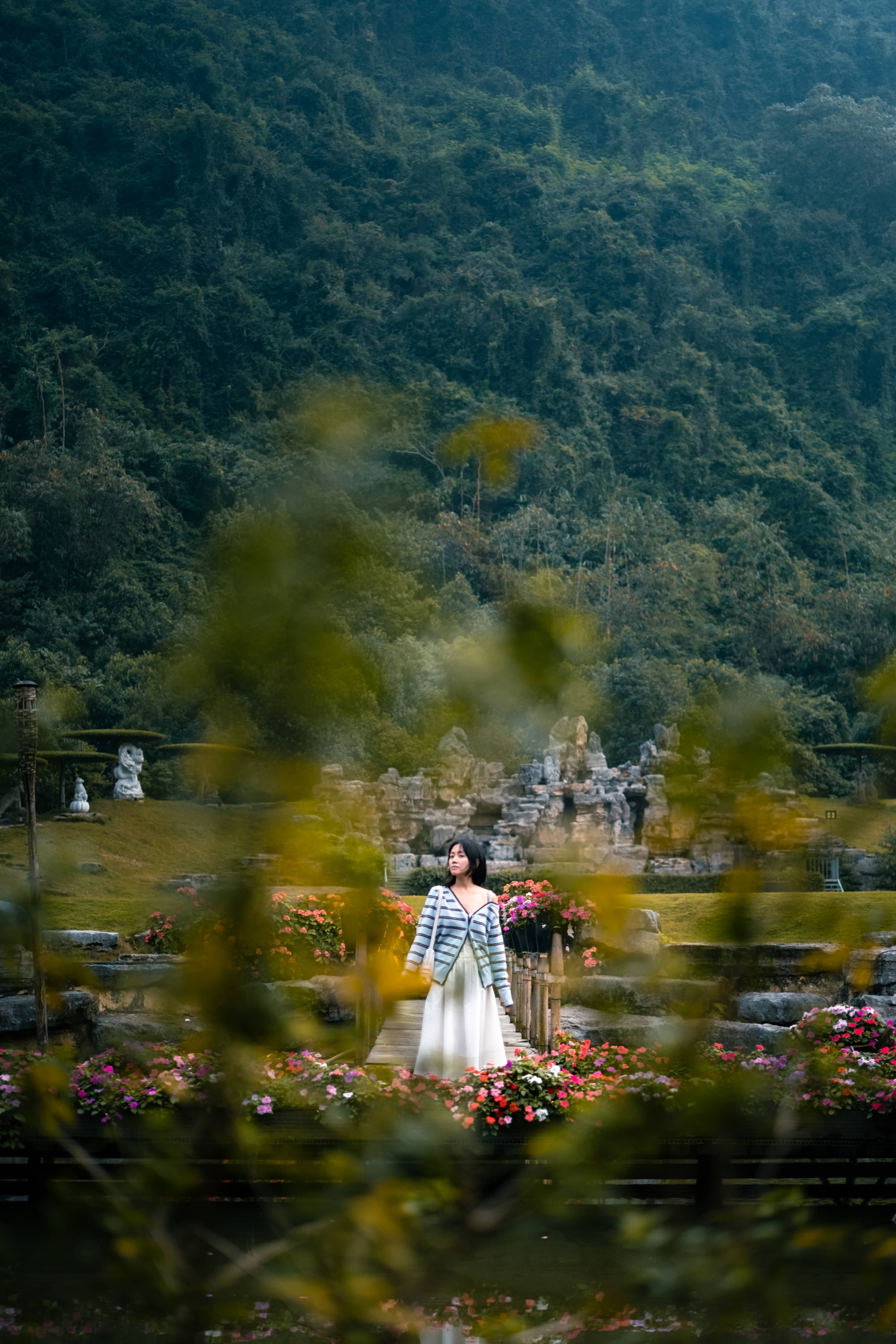Woman in a striped top and white skirt framed by blurred foliage in a garden below forested hills