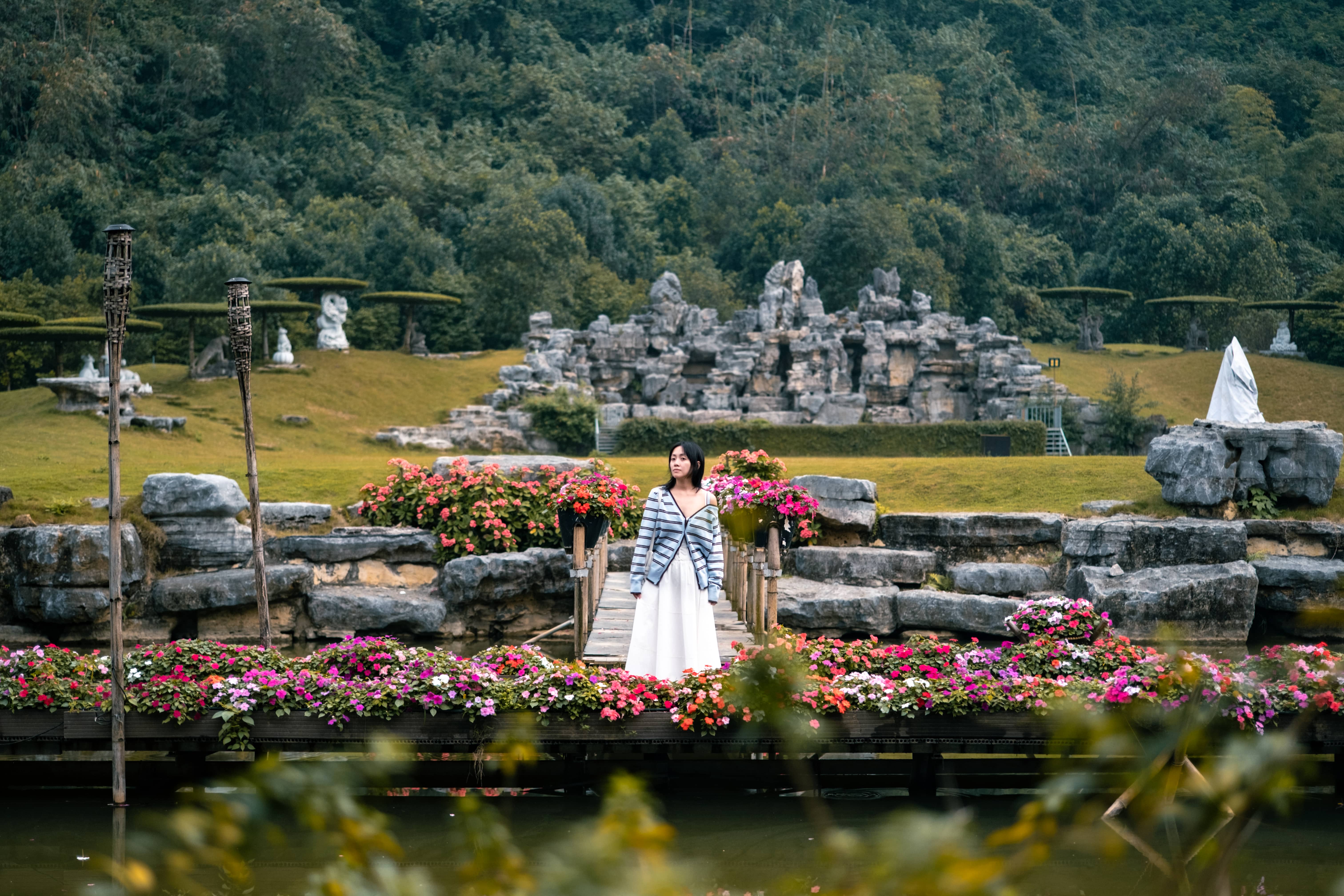 Woman in a striped top on a small bridge amid pink flowers, a rock garden and forest behind, Vietnam