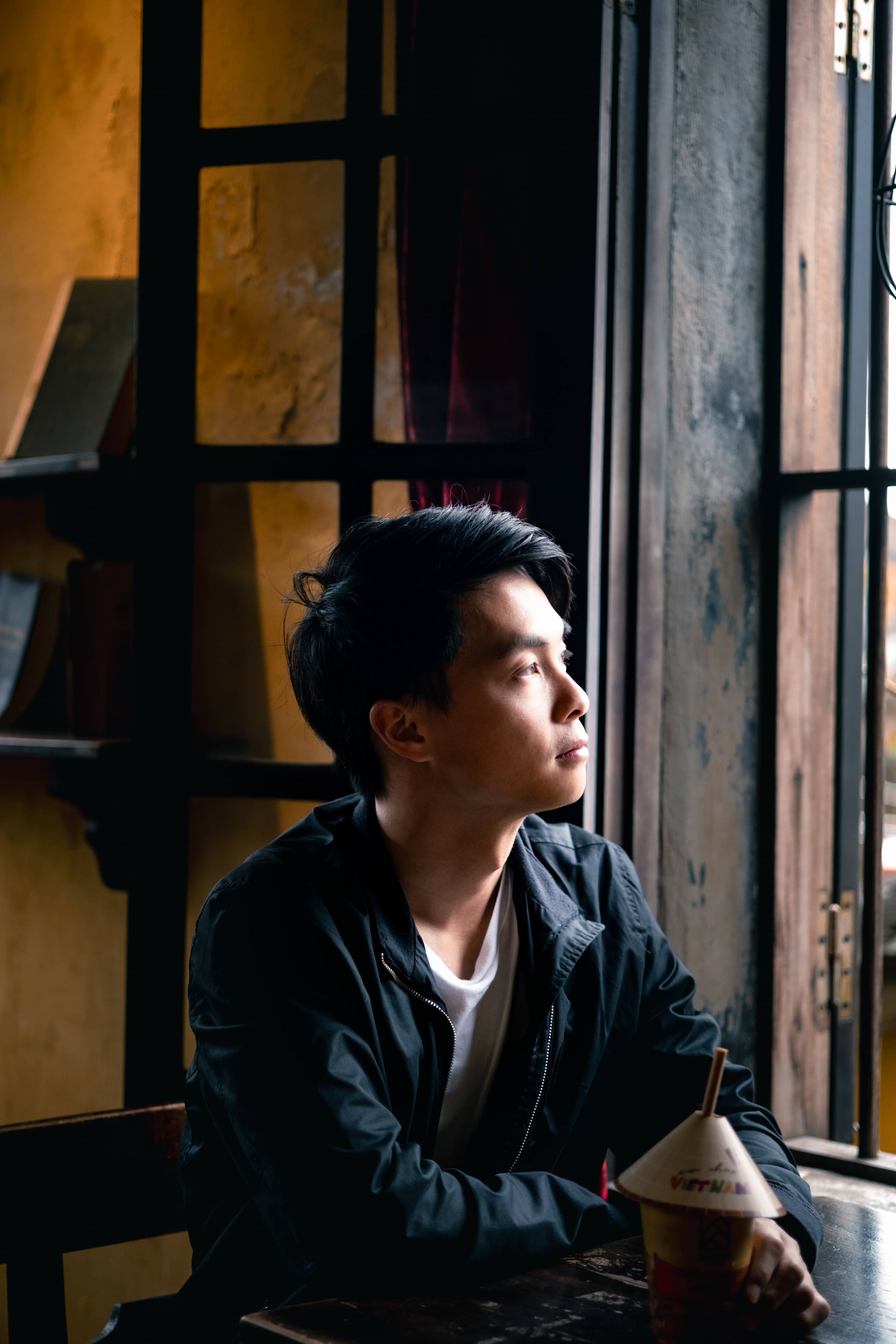 Man in a black jacket sitting by a window in a warm-lit cafe, looking out, a conical-hat cup on the table, Vietnam