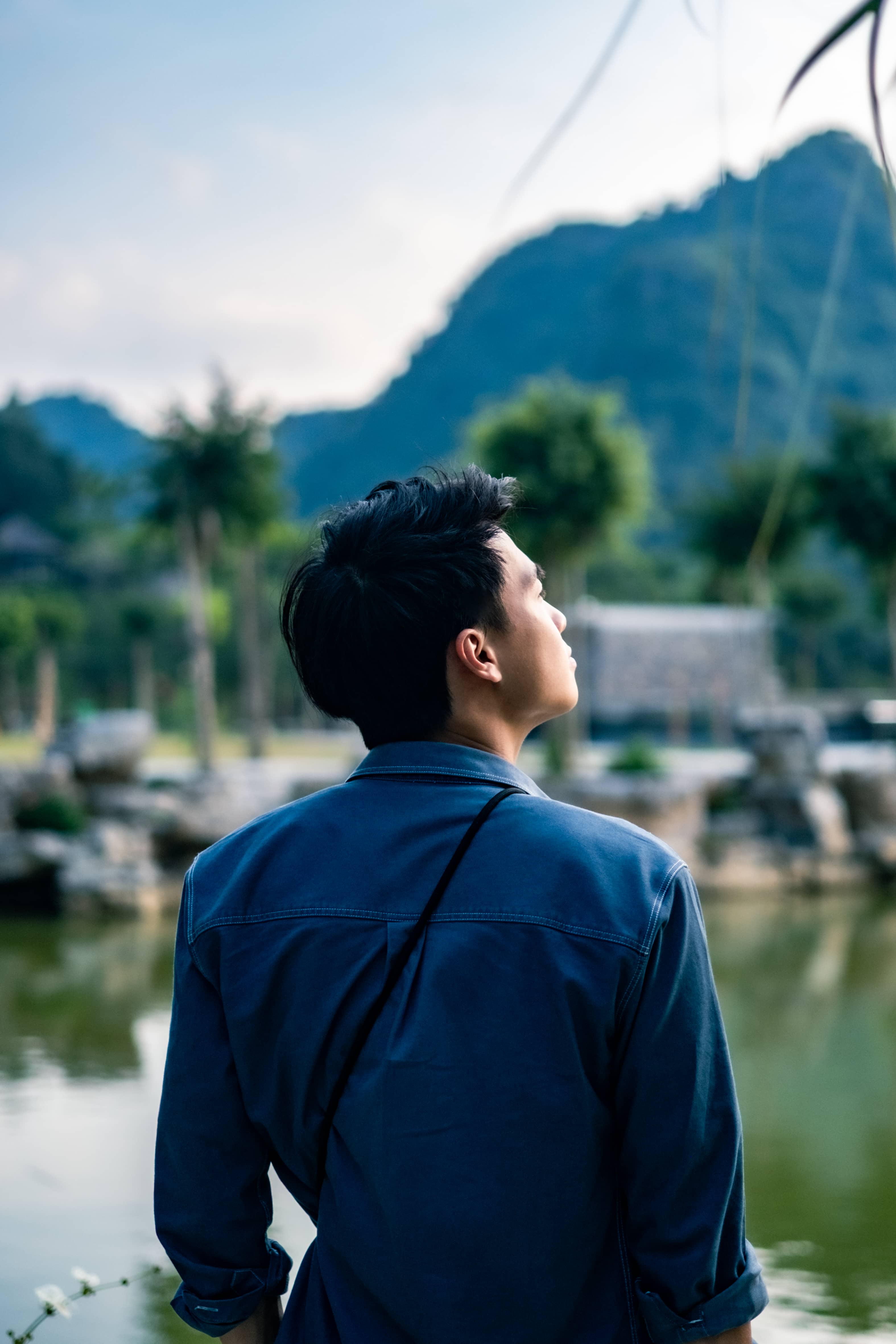 Man in a denim shirt seen from behind, looking across a lake toward green limestone hills, Vietnam