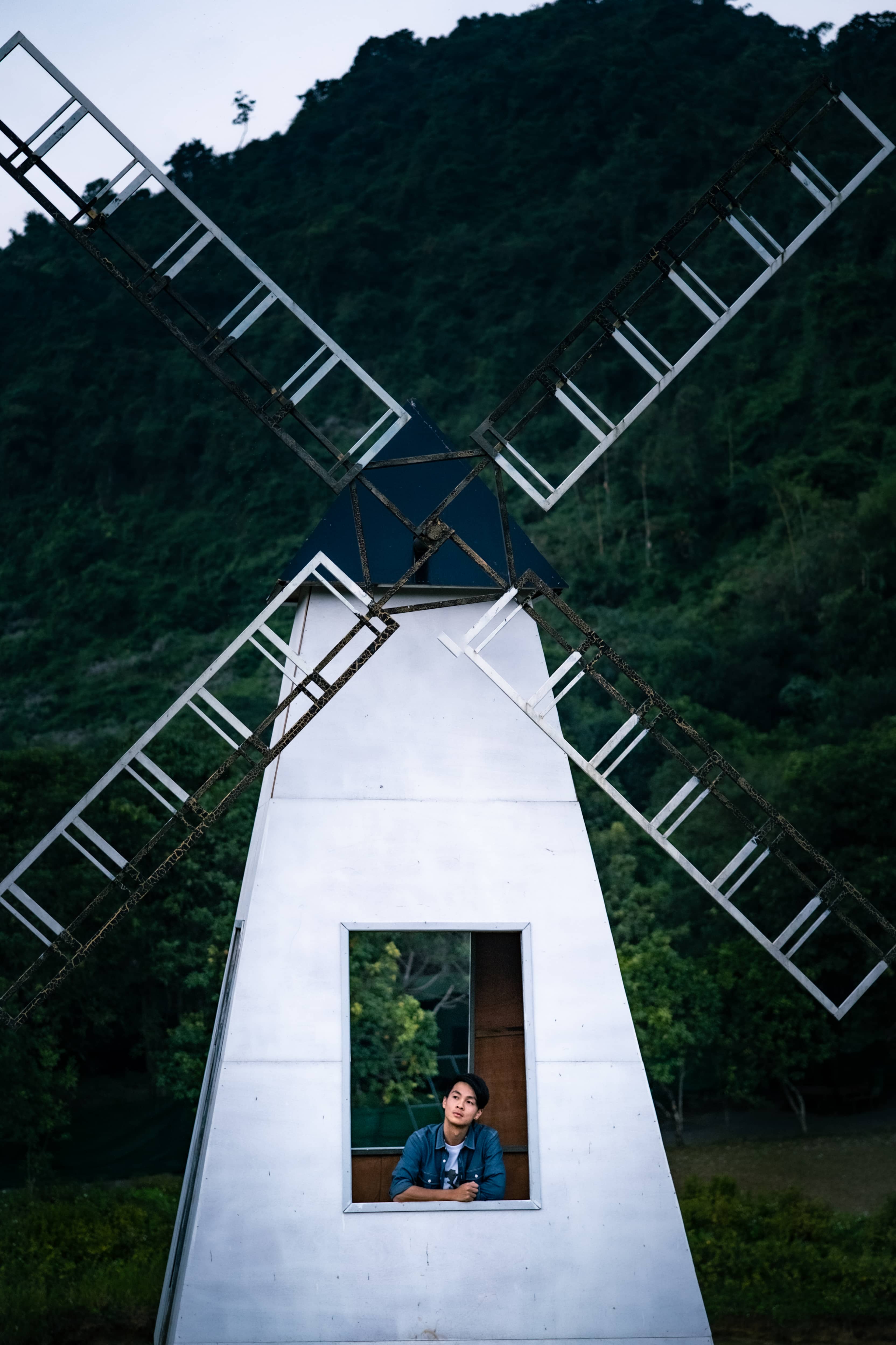 Man in a denim jacket looking out from the window of a white windmill, green forested hill behind