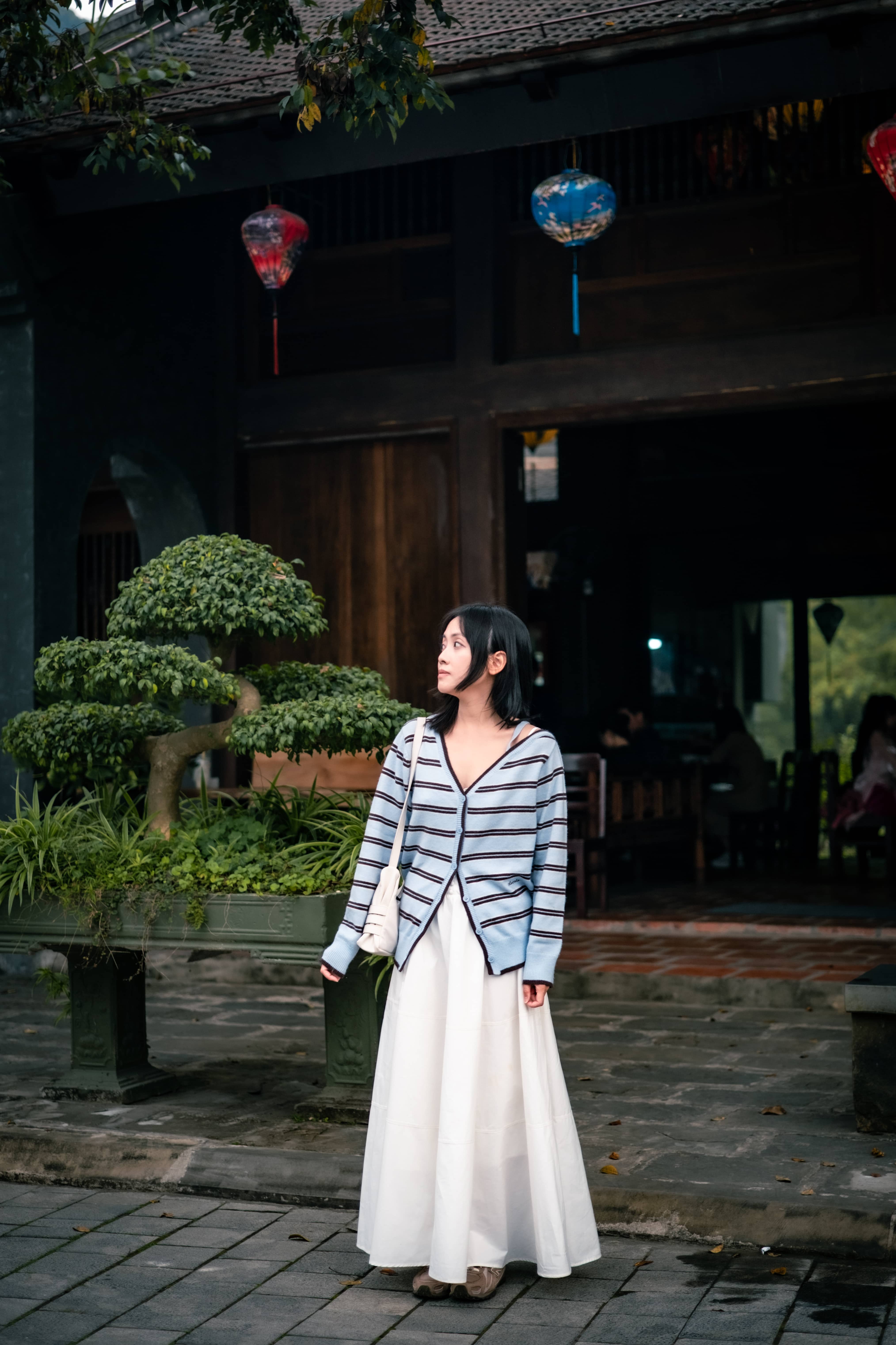 Woman in a striped cardigan and long white skirt standing before a dark wooden building with colourful lanterns and a bonsai tree, Vietnam