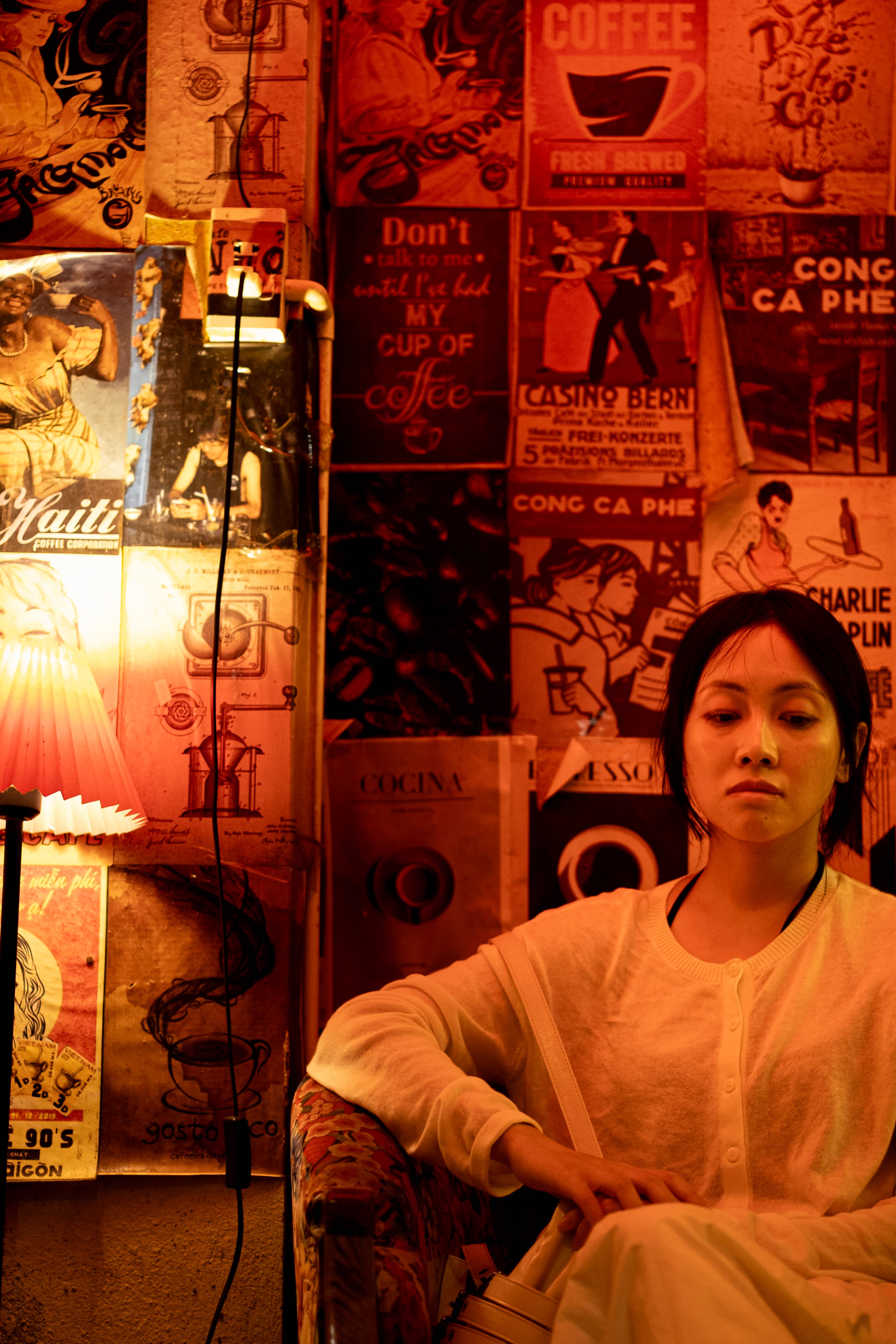 Woman in a cream cardigan seated in a red-lit cafe, gazing down, vintage coffee posters behind, Vietnam