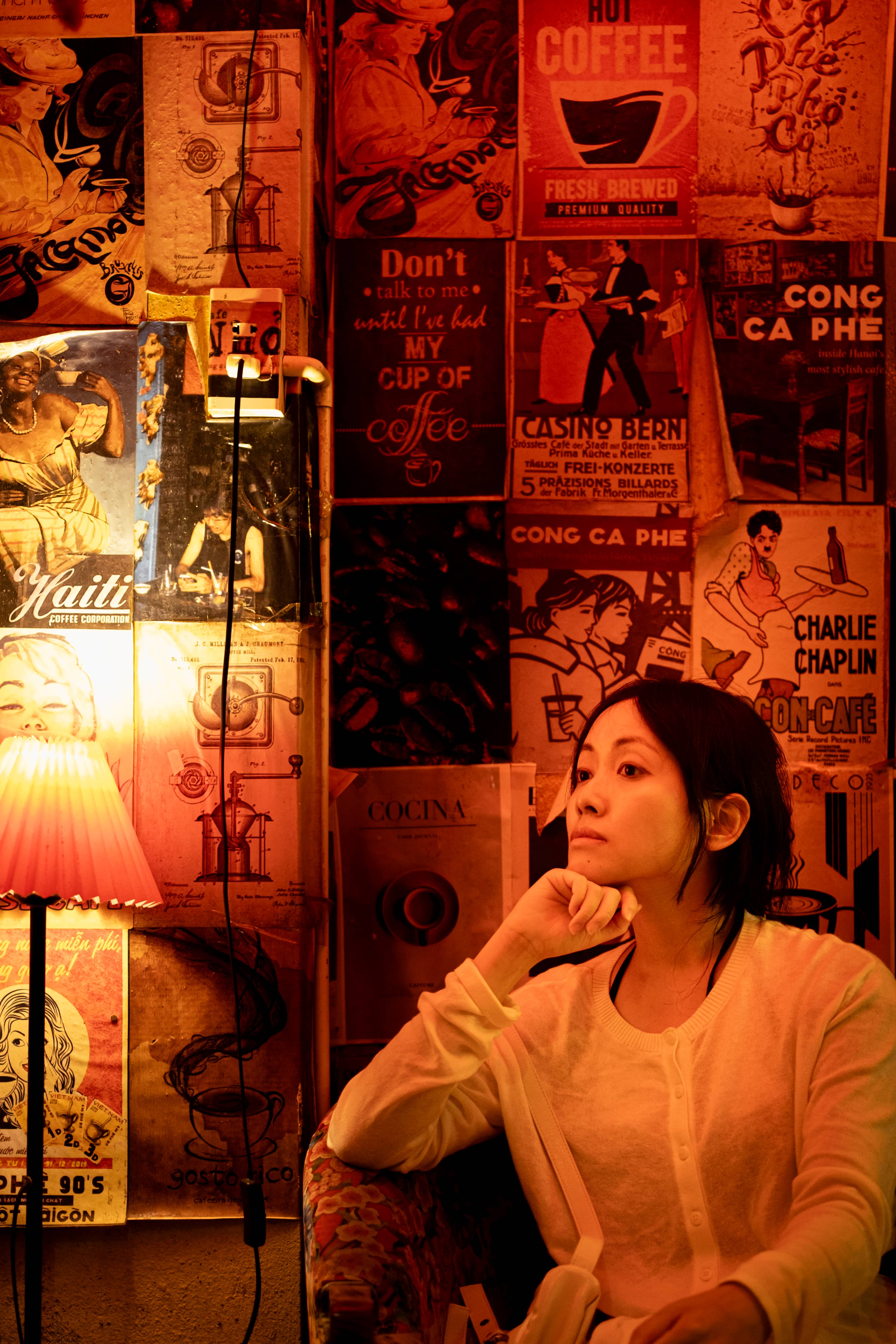 Woman resting her chin on her hand in a warm red-lit cafe, walls covered in vintage coffee posters, Vietnam