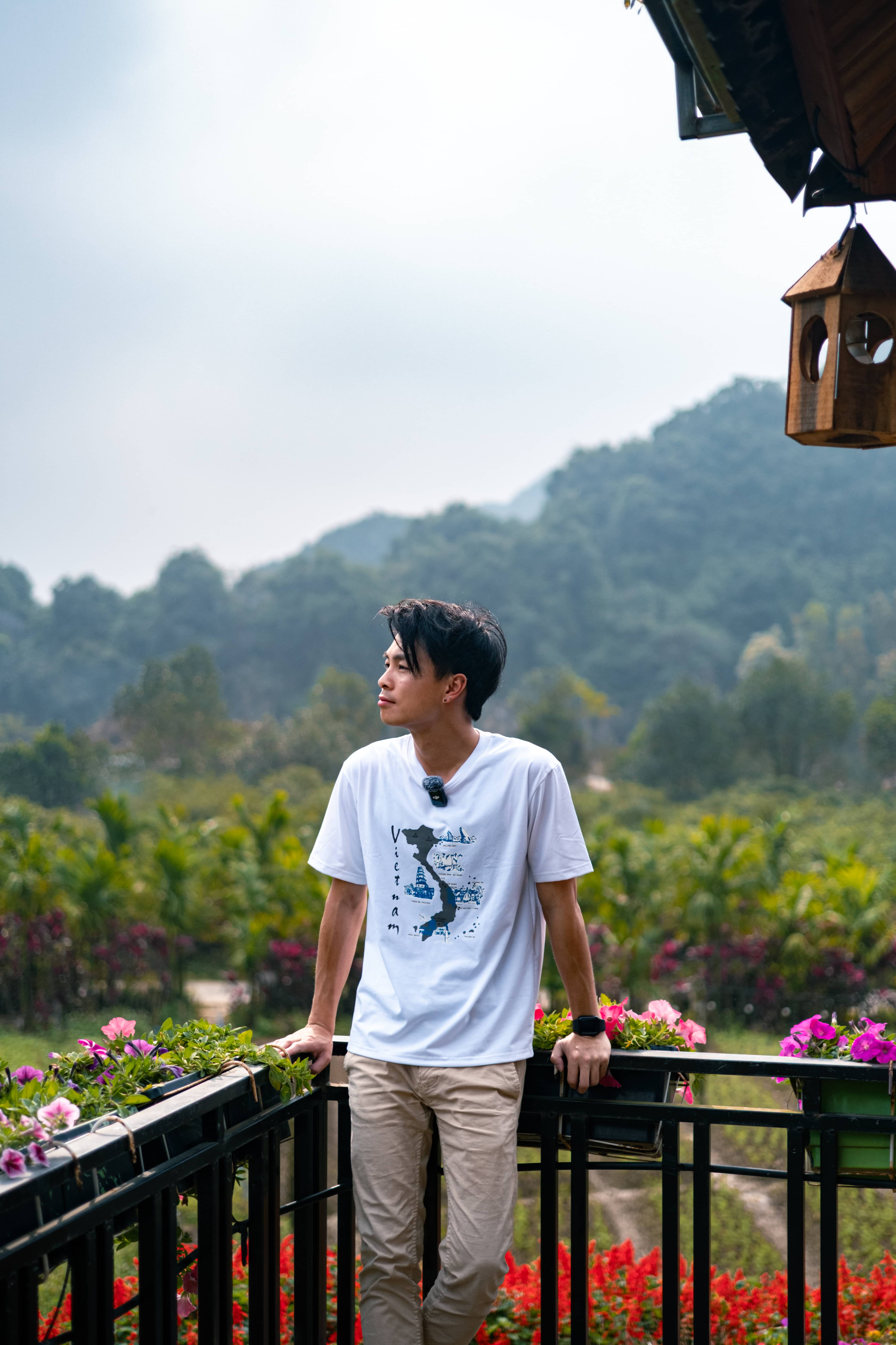 Man in a white Vietnam map t-shirt leaning on a balcony rail with pink flowers, misty green hills behind