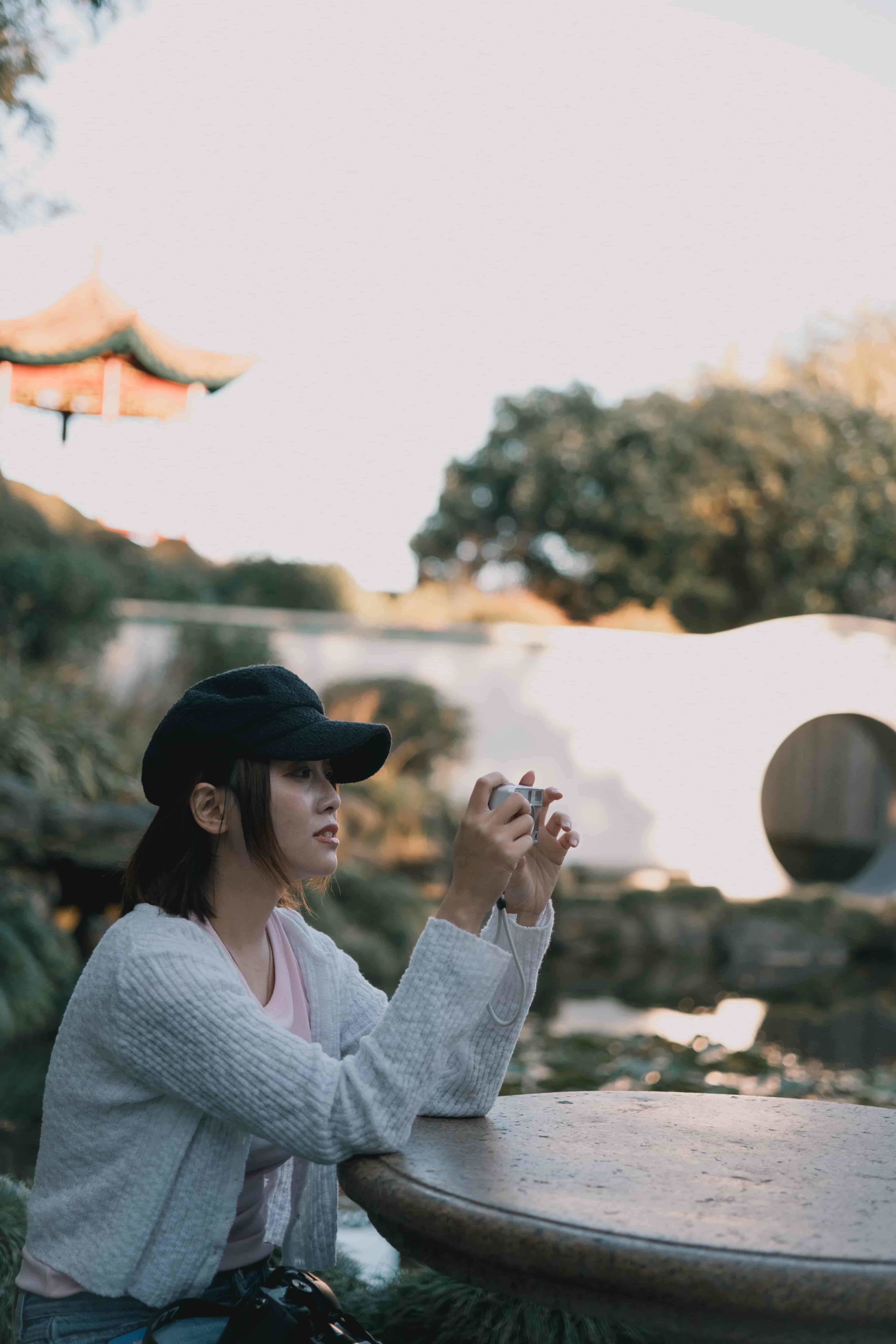 Woman in a cap and cardigan framing a shot with a small camera beside a pond in a Chinese-style garden