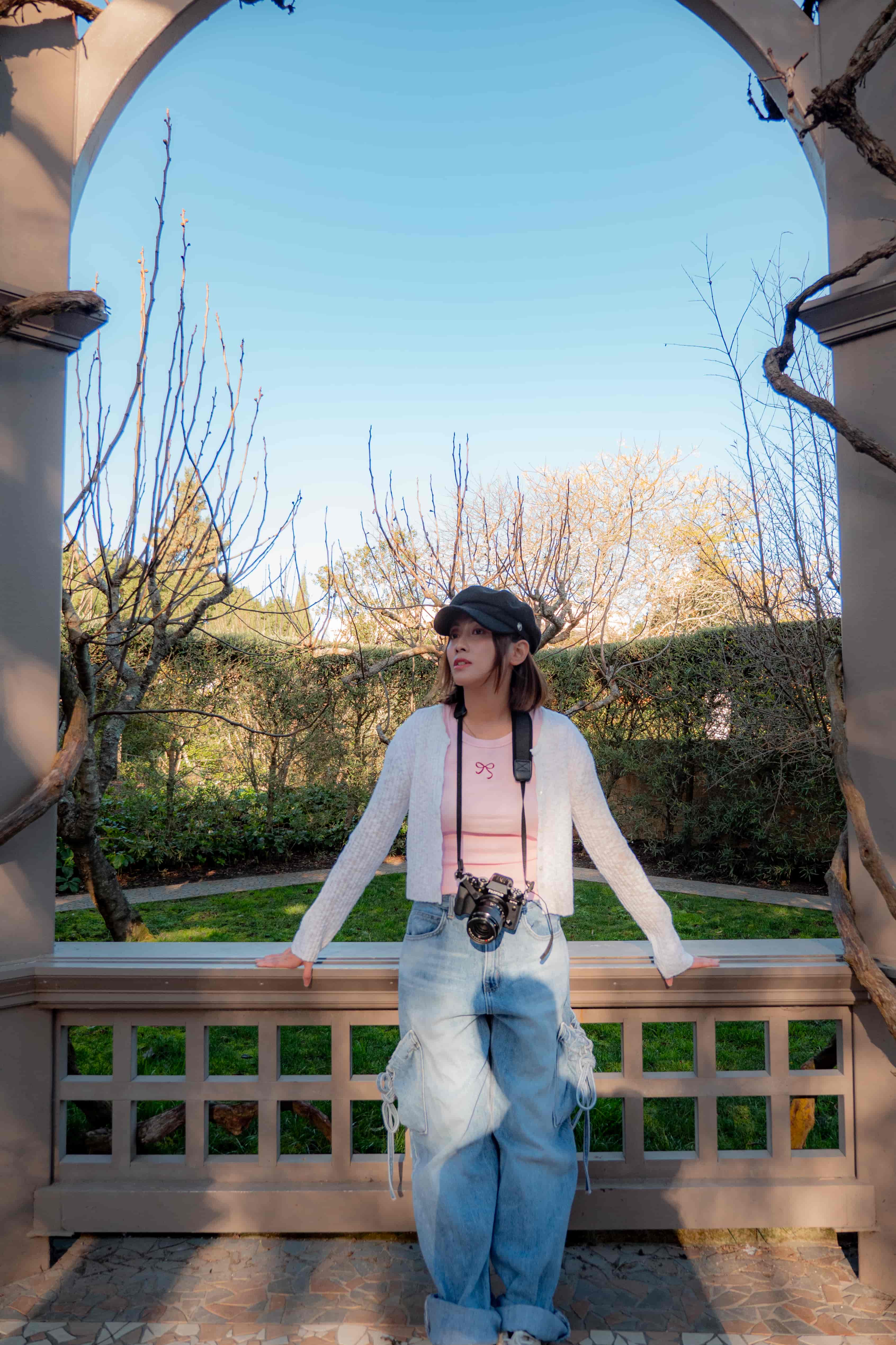 Woman in a cap and wide jeans leaning on a timber rail under a vine-covered arbour, blue sky above