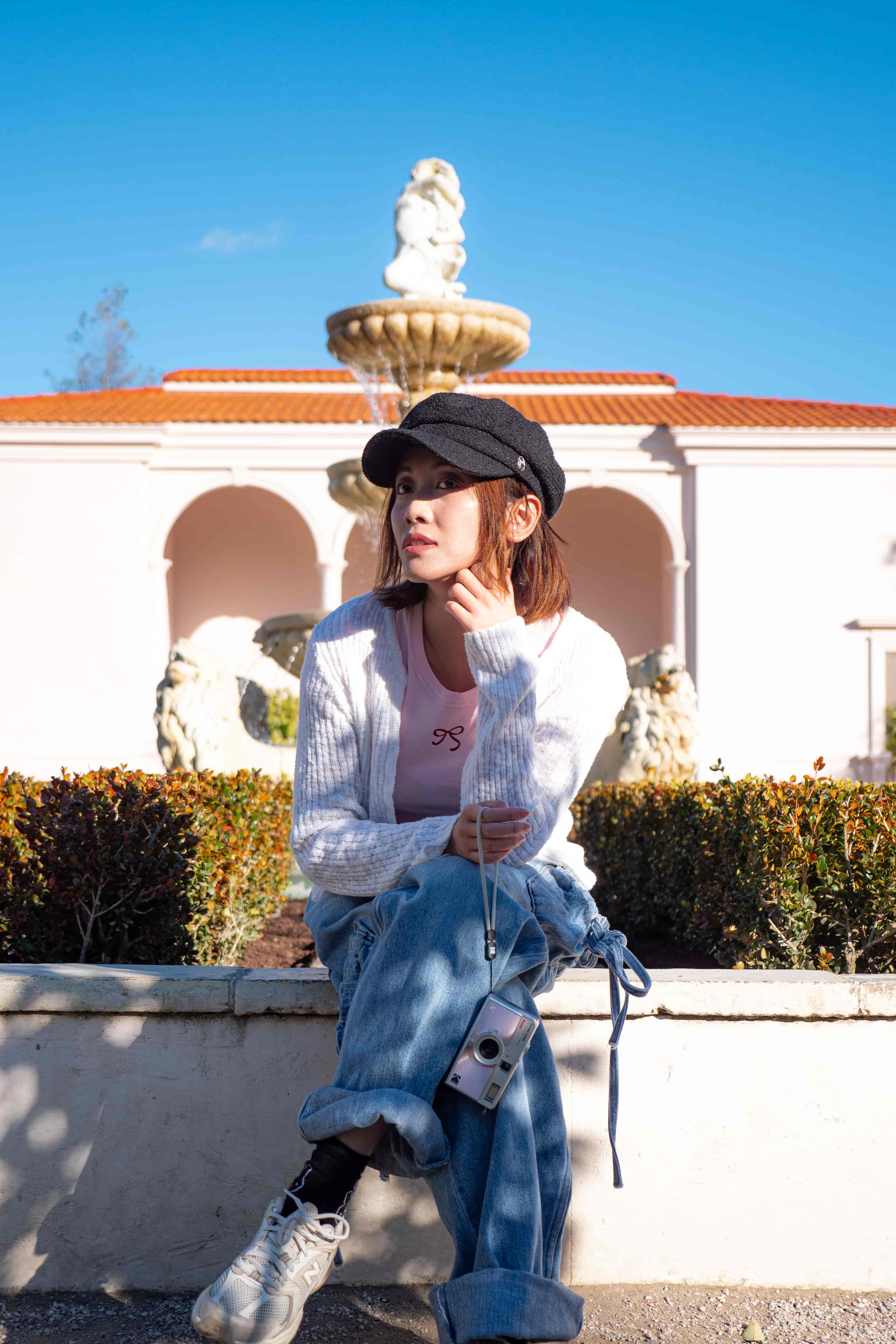 Woman in a cap seated on a low wall before a white villa with arches and a tiered fountain