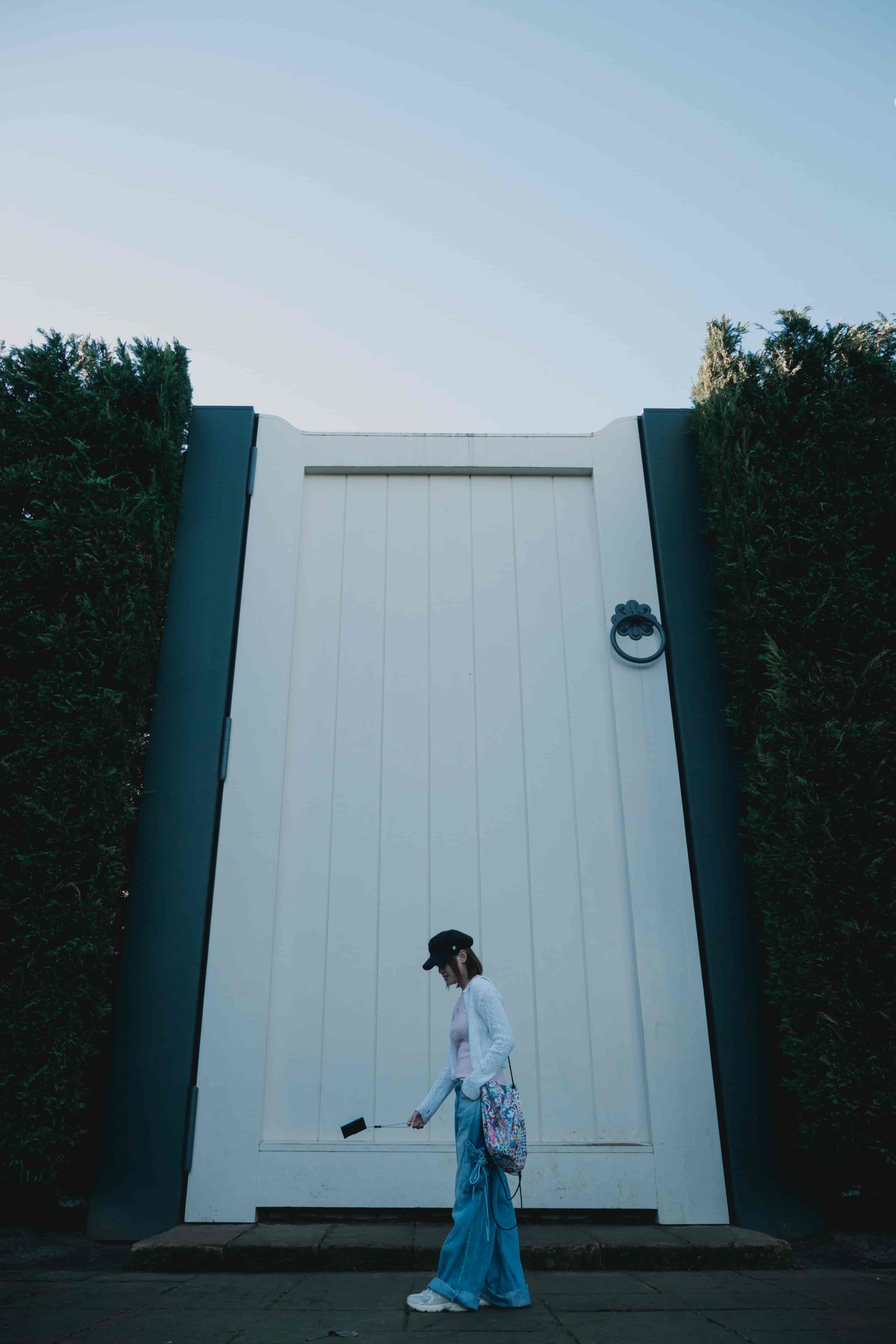 Woman walking past a tall white garden door set between dark clipped hedges, holding a small camera