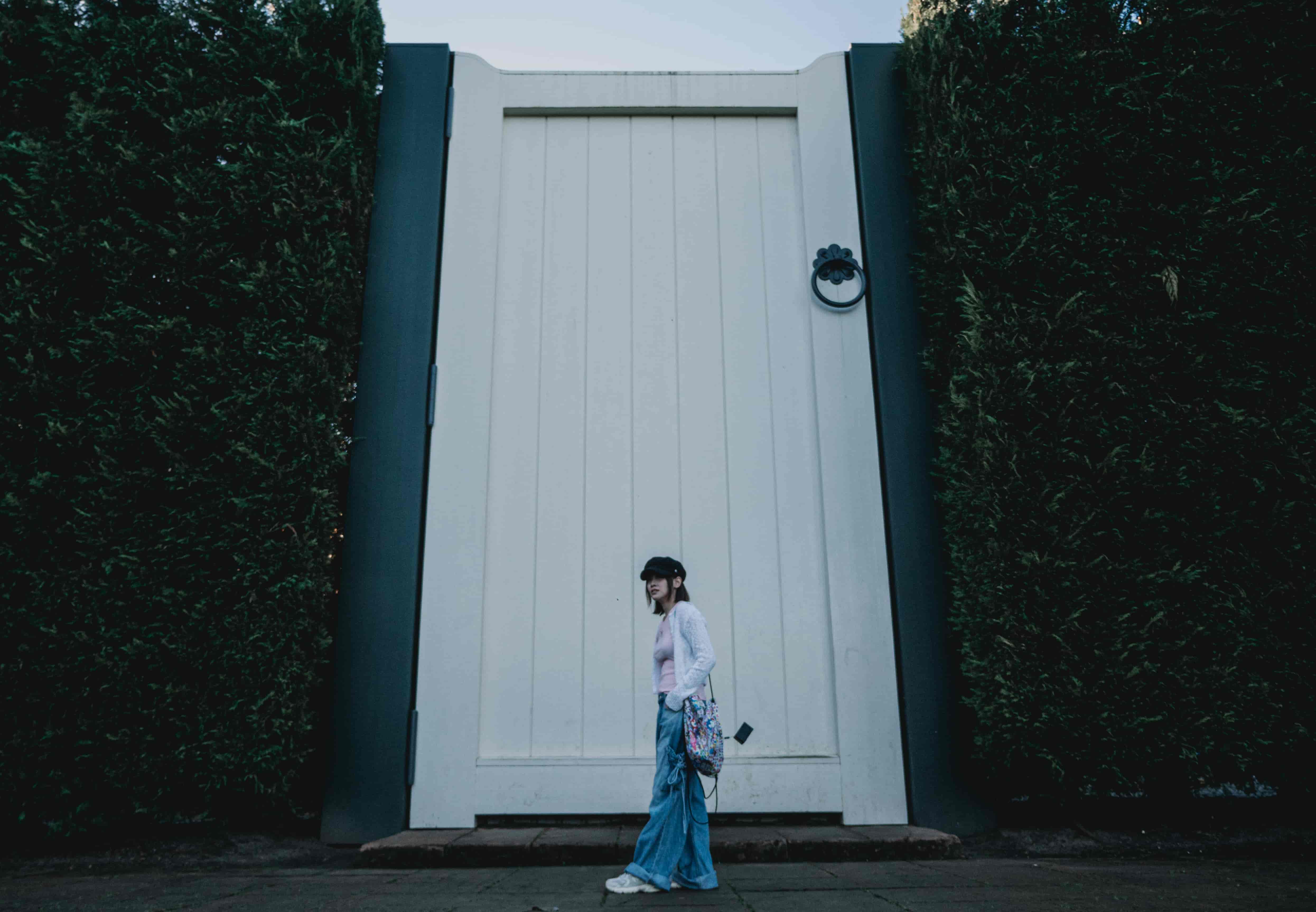 Wide view of a woman walking before an oversized white door framed by tall dark hedges