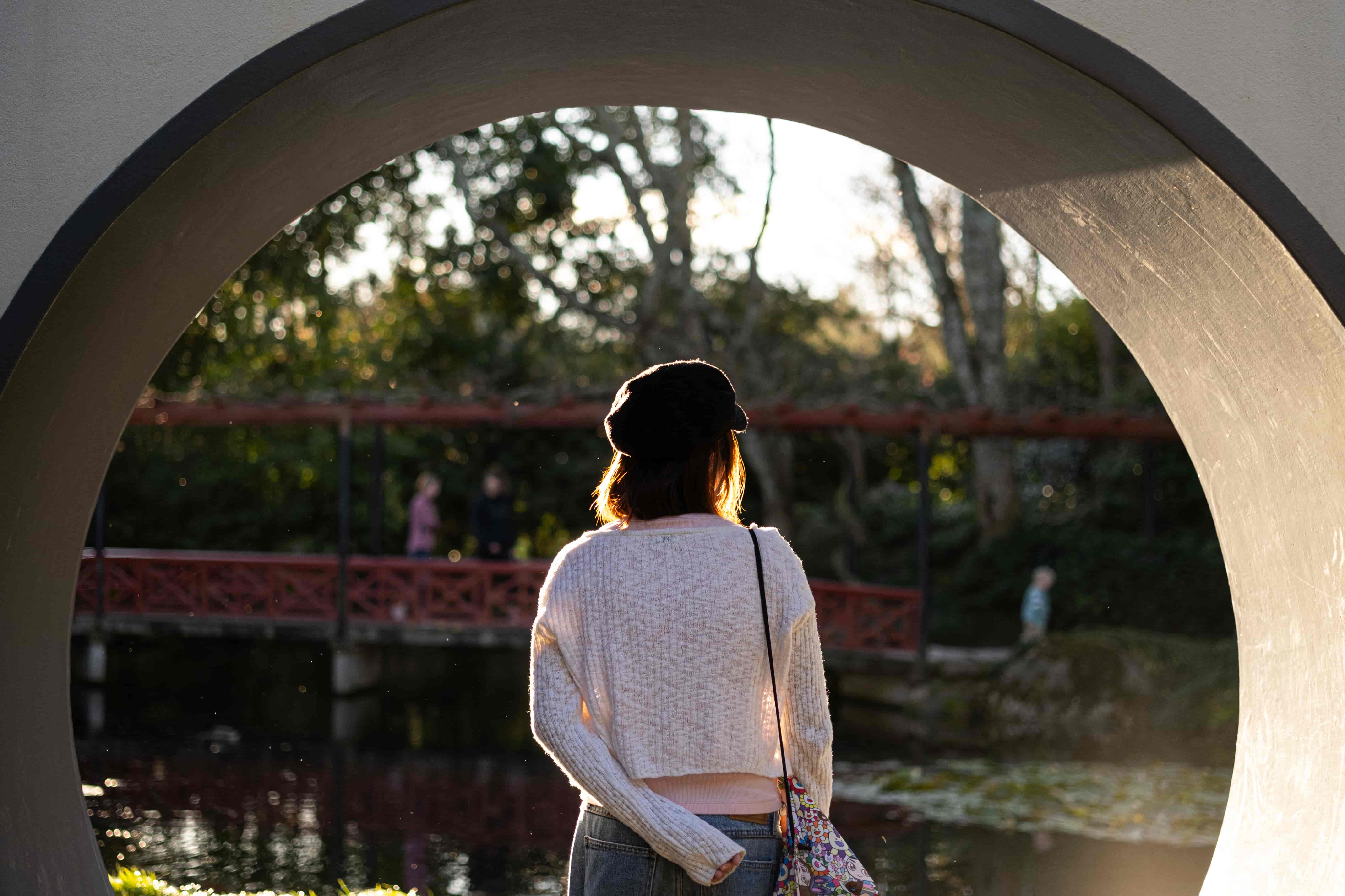 Woman seen from behind through a round moon gate, looking toward a pond and a red bridge in late light
