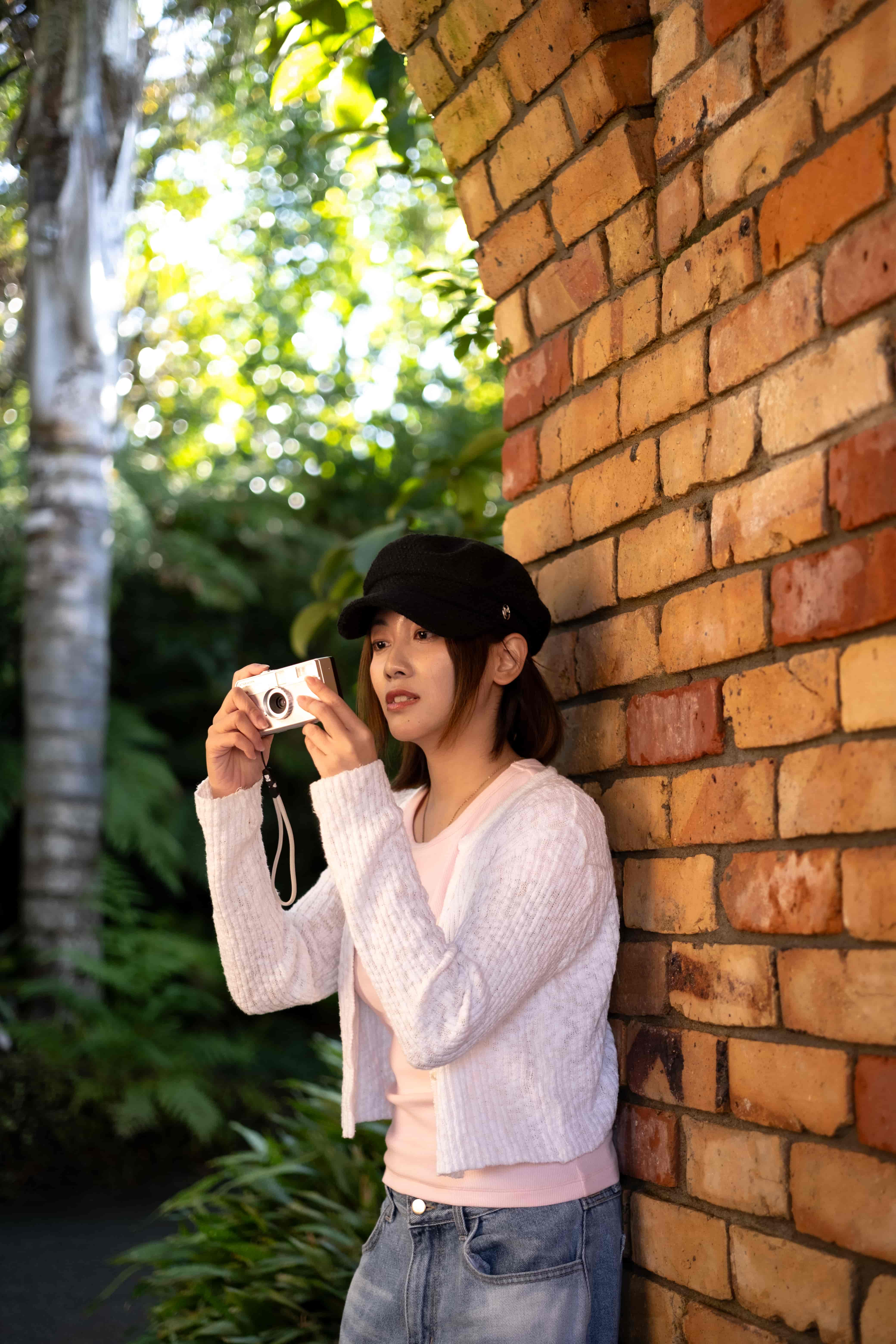 Woman holding a compact camera against a red brick wall, with ferns and trees glowing in sunlight