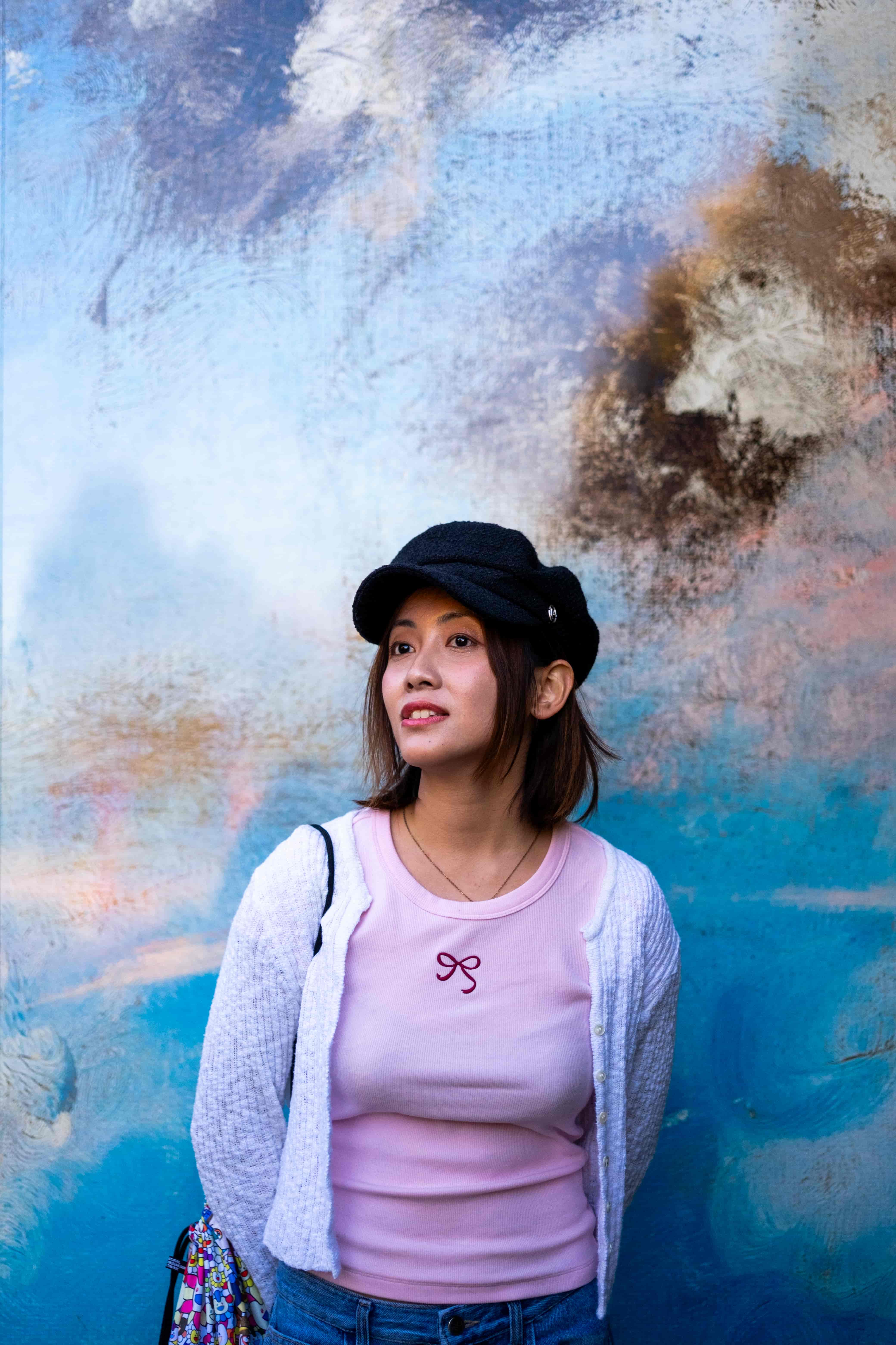 Woman in a cap and pink top leaning against a weathered painted wall in blue, white and rust tones
