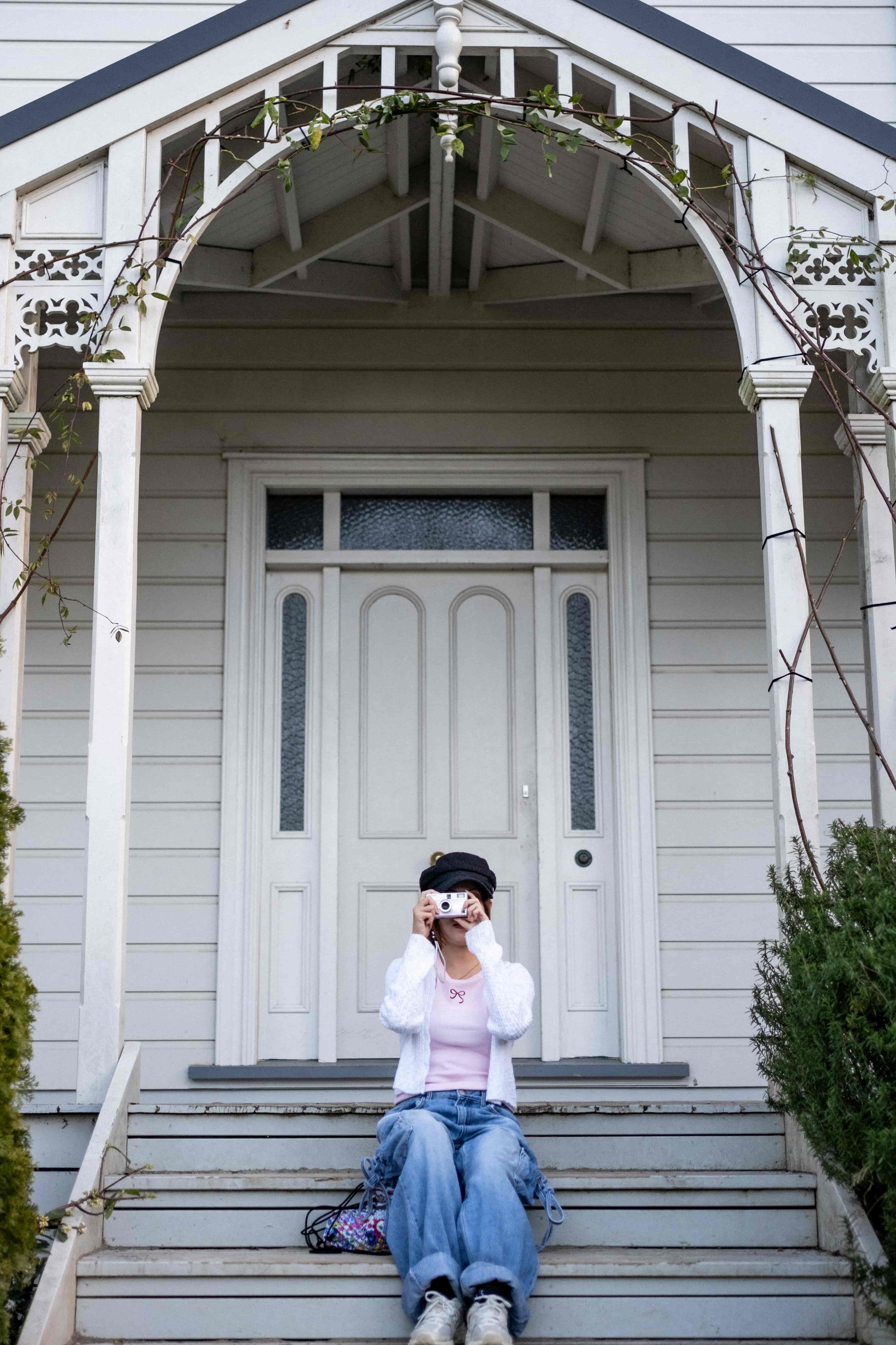 Woman on the steps of a white villa porch photographing with a compact camera held to her eye