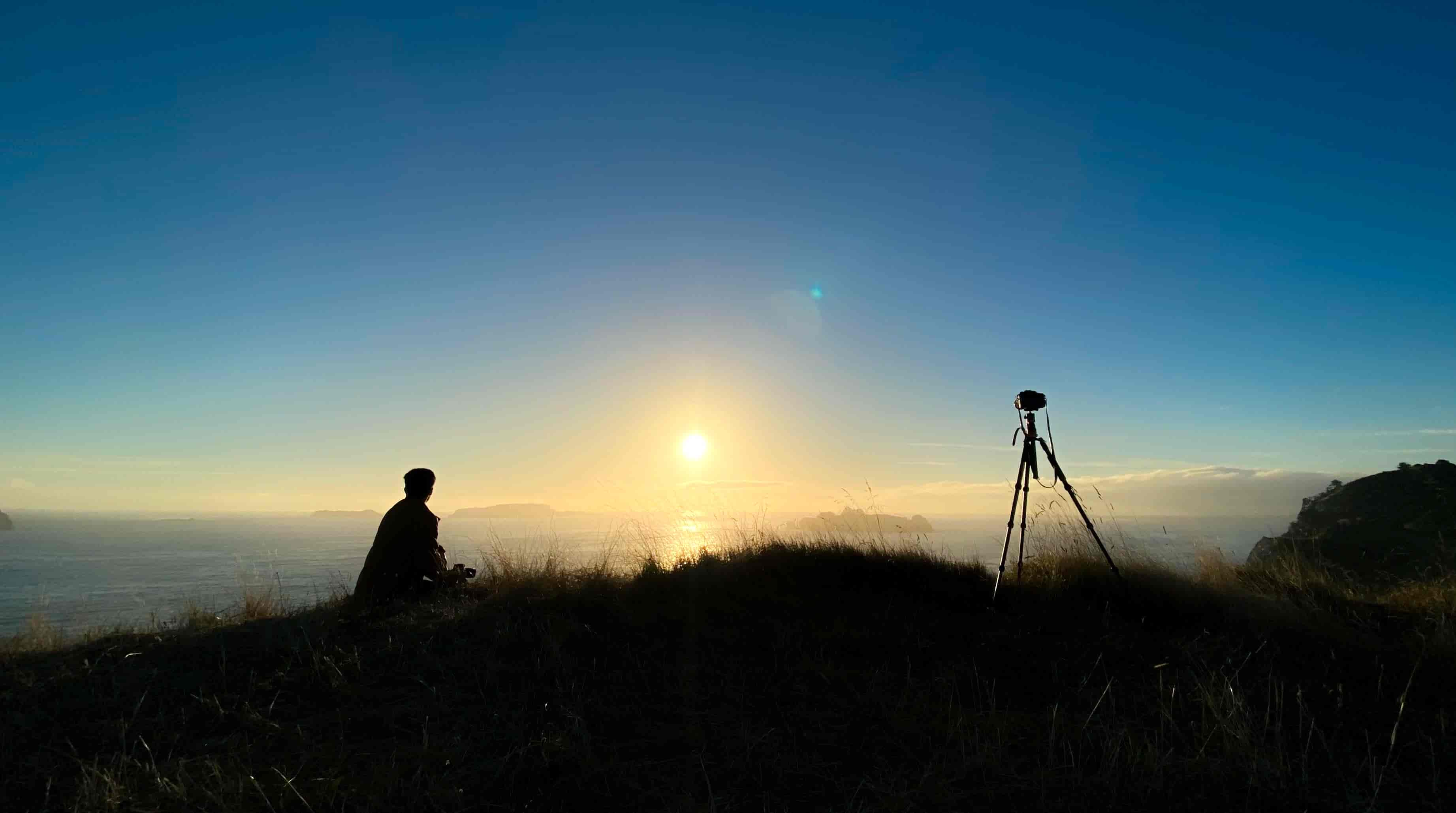Silhouetted person sitting beside a tripod on a grassy clifftop as the sun sets over the sea