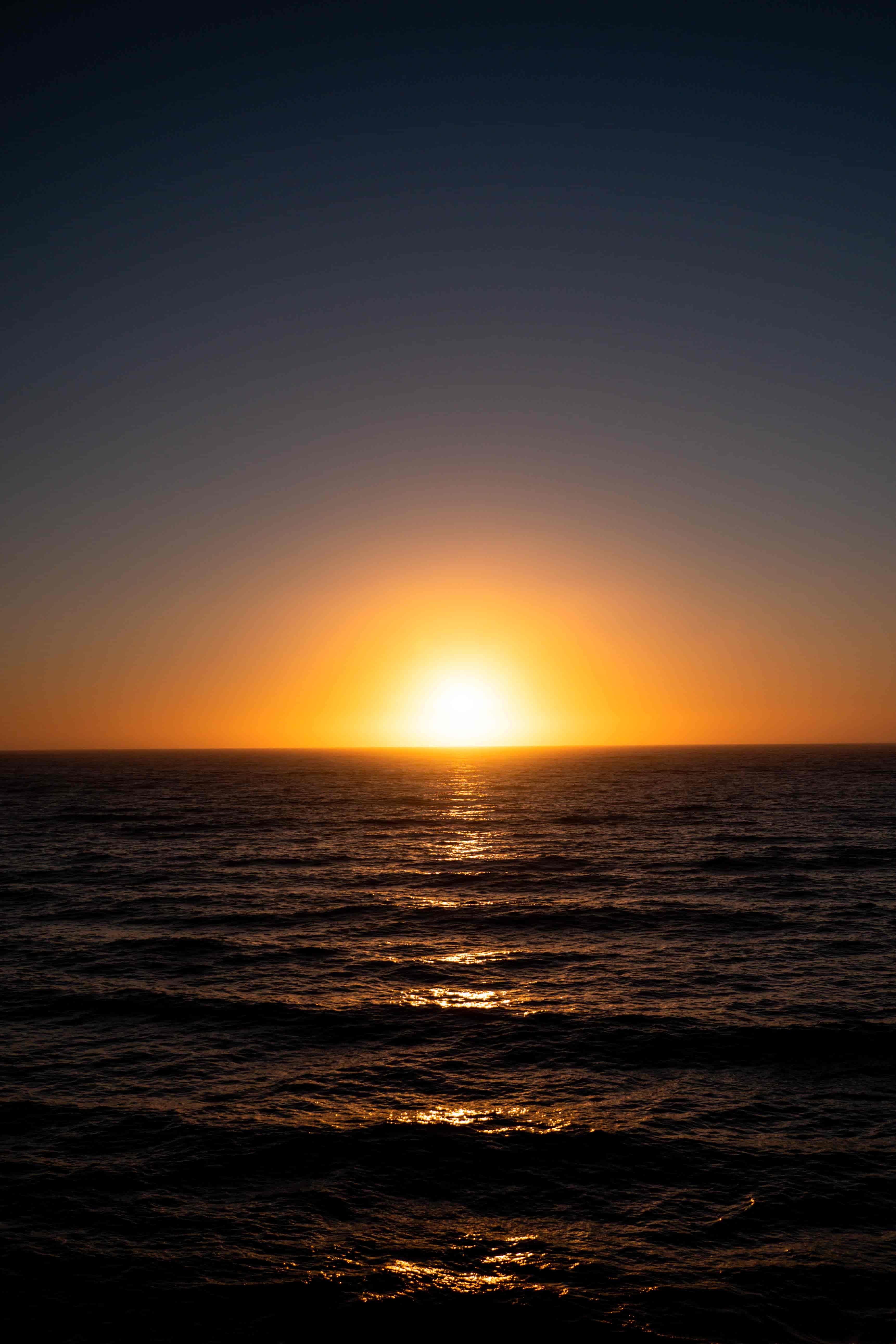 Sun setting into a calm dark sea, a golden glow spreading across the horizon and the water