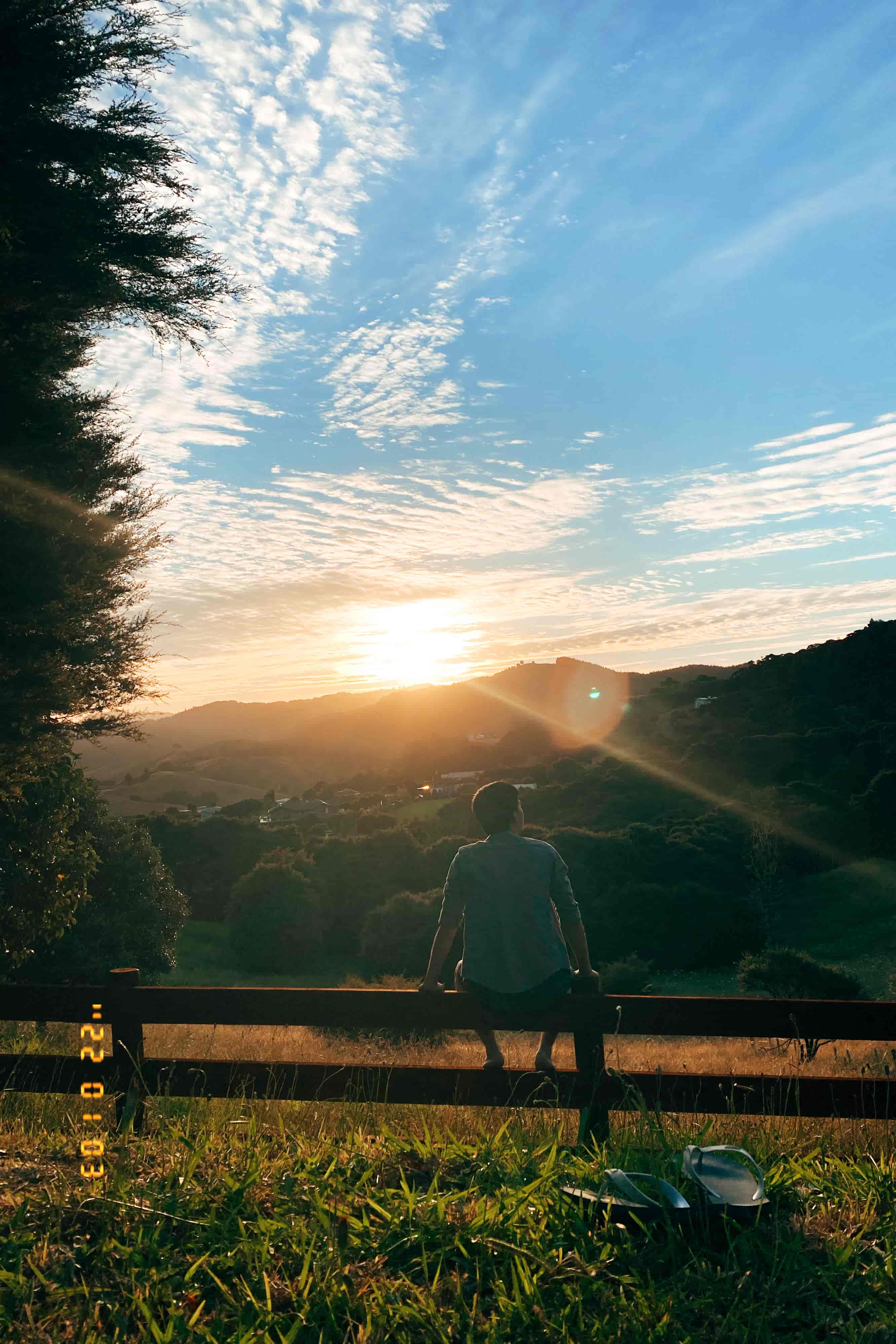 Person sitting on a wooden fence watching the sun set over green hills beneath streaky clouds