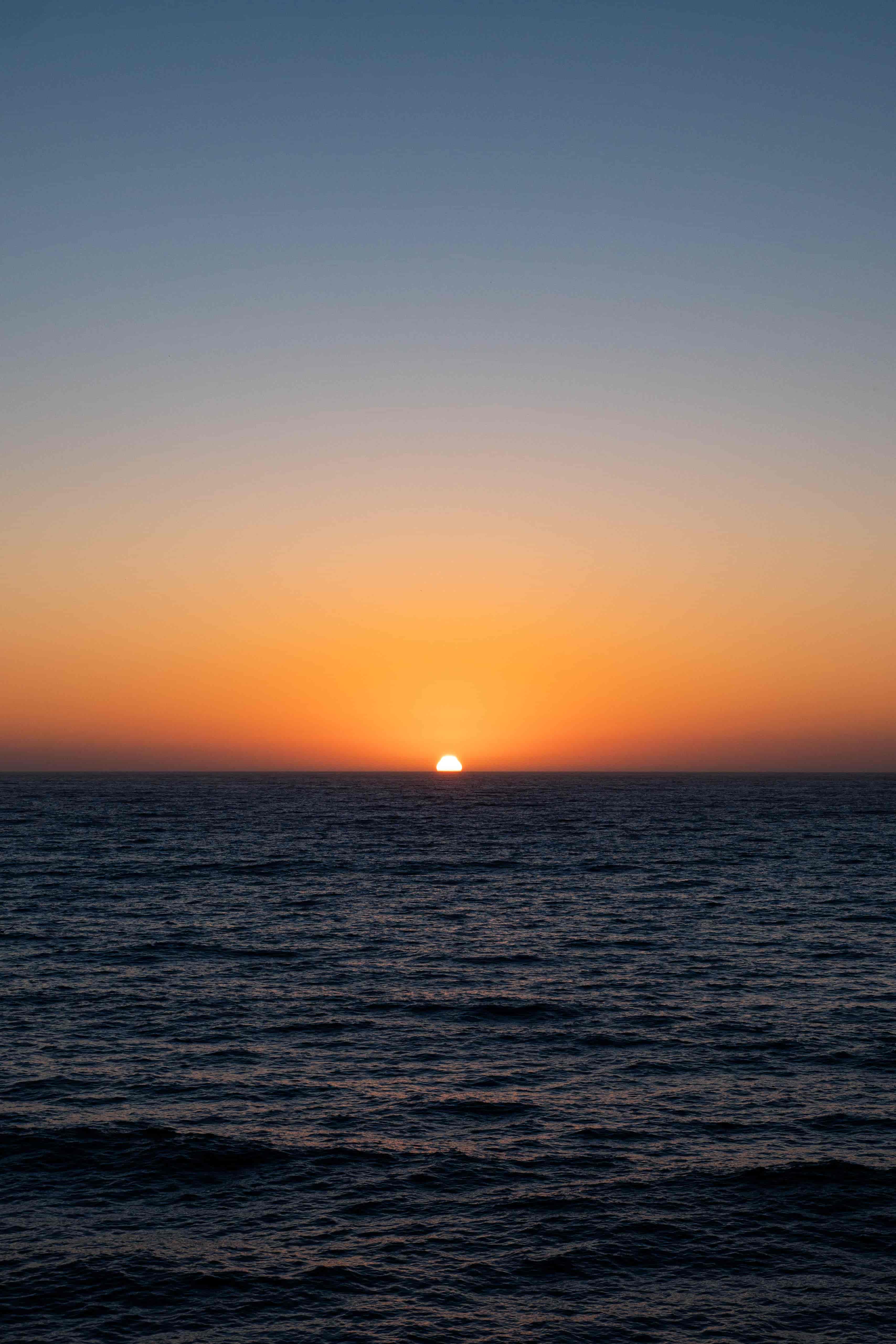 Last sliver of the sun dipping below a flat ocean horizon in an orange and blue dusk sky