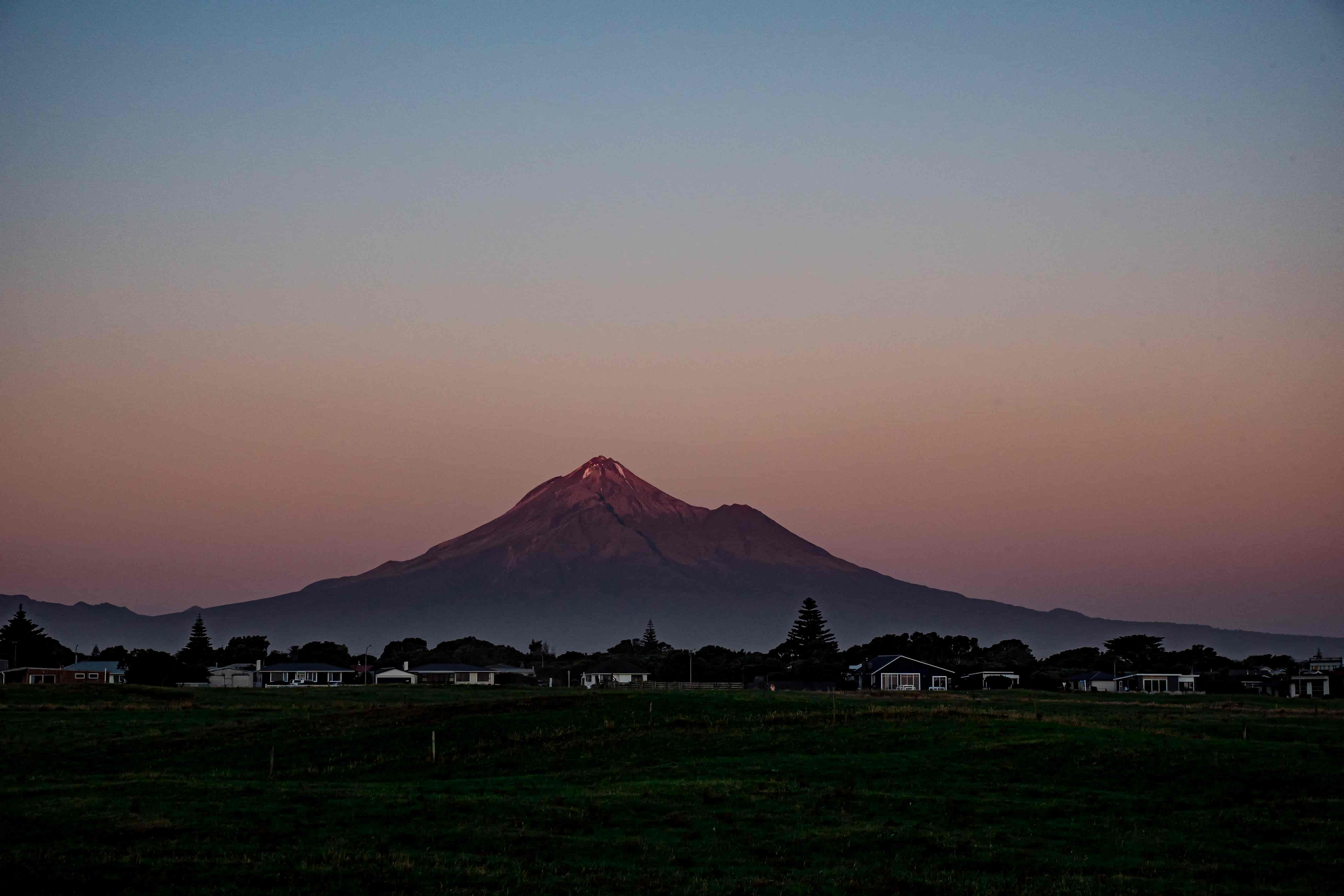Mount Taranaki glowing pink at dusk above dark farmland and a row of houses