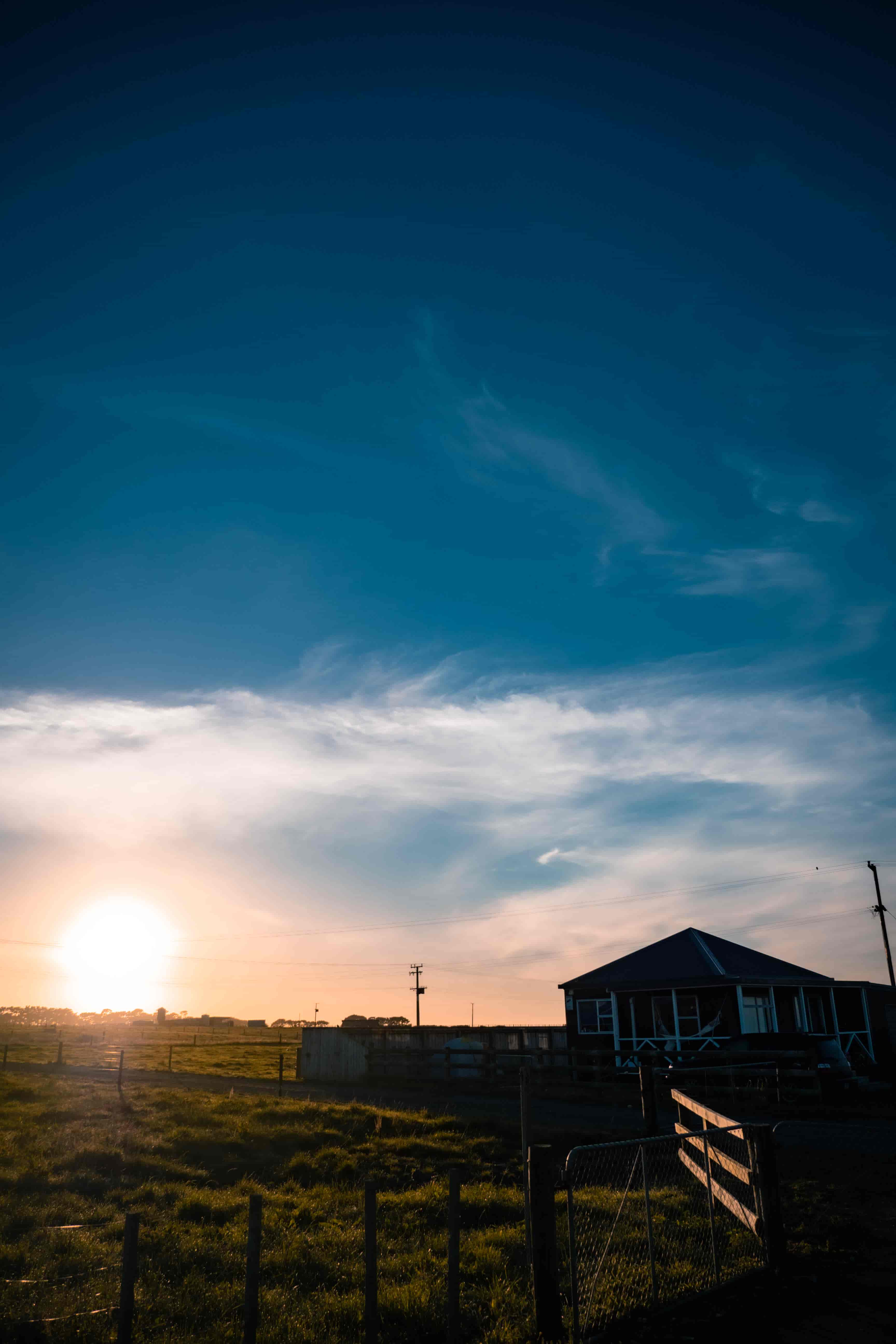 Sun setting low behind a farm shed and wooden fence under a deep blue sky with wispy cloud