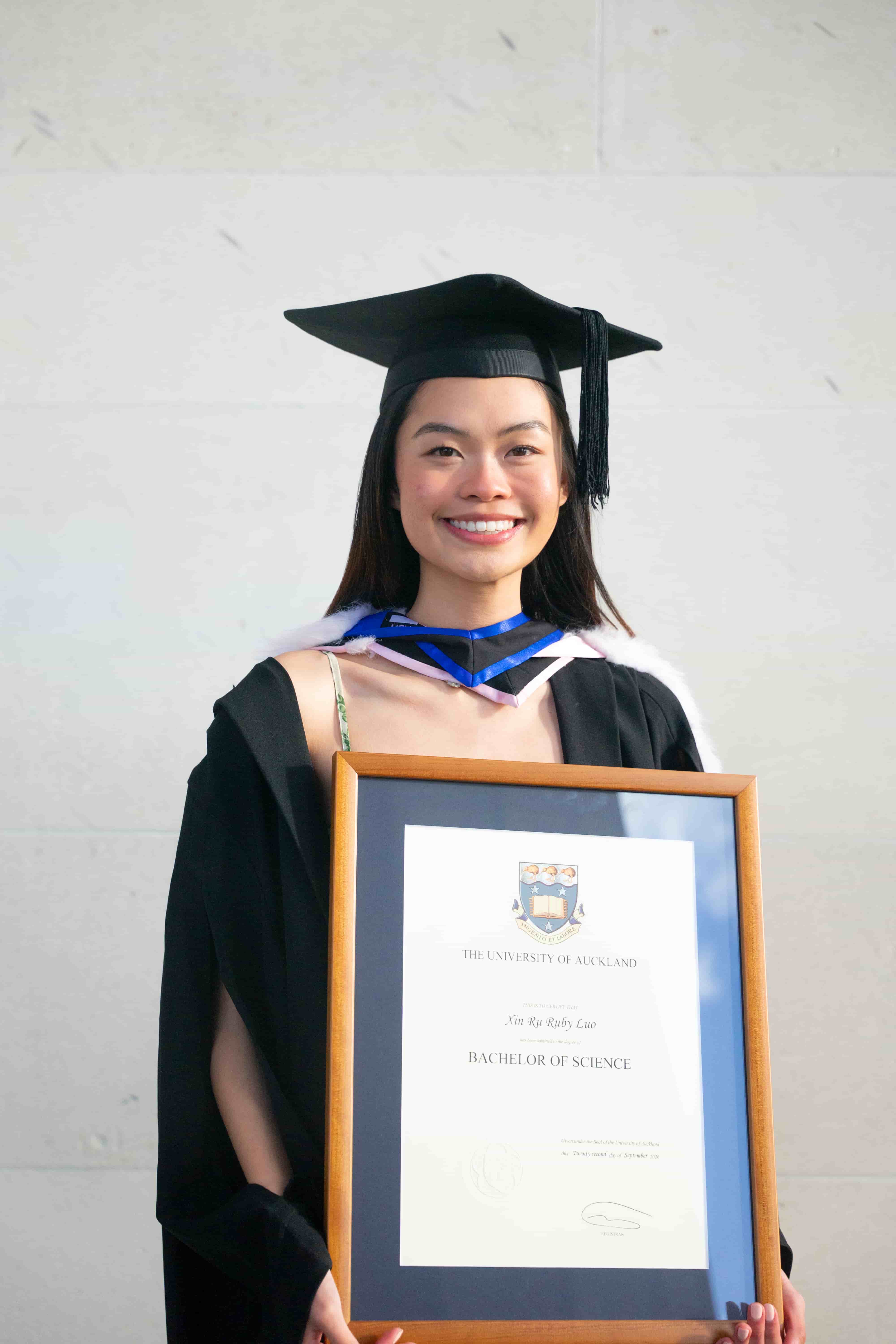 Ruby in graduation cap and gown smiling with her framed Bachelor of Science certificate