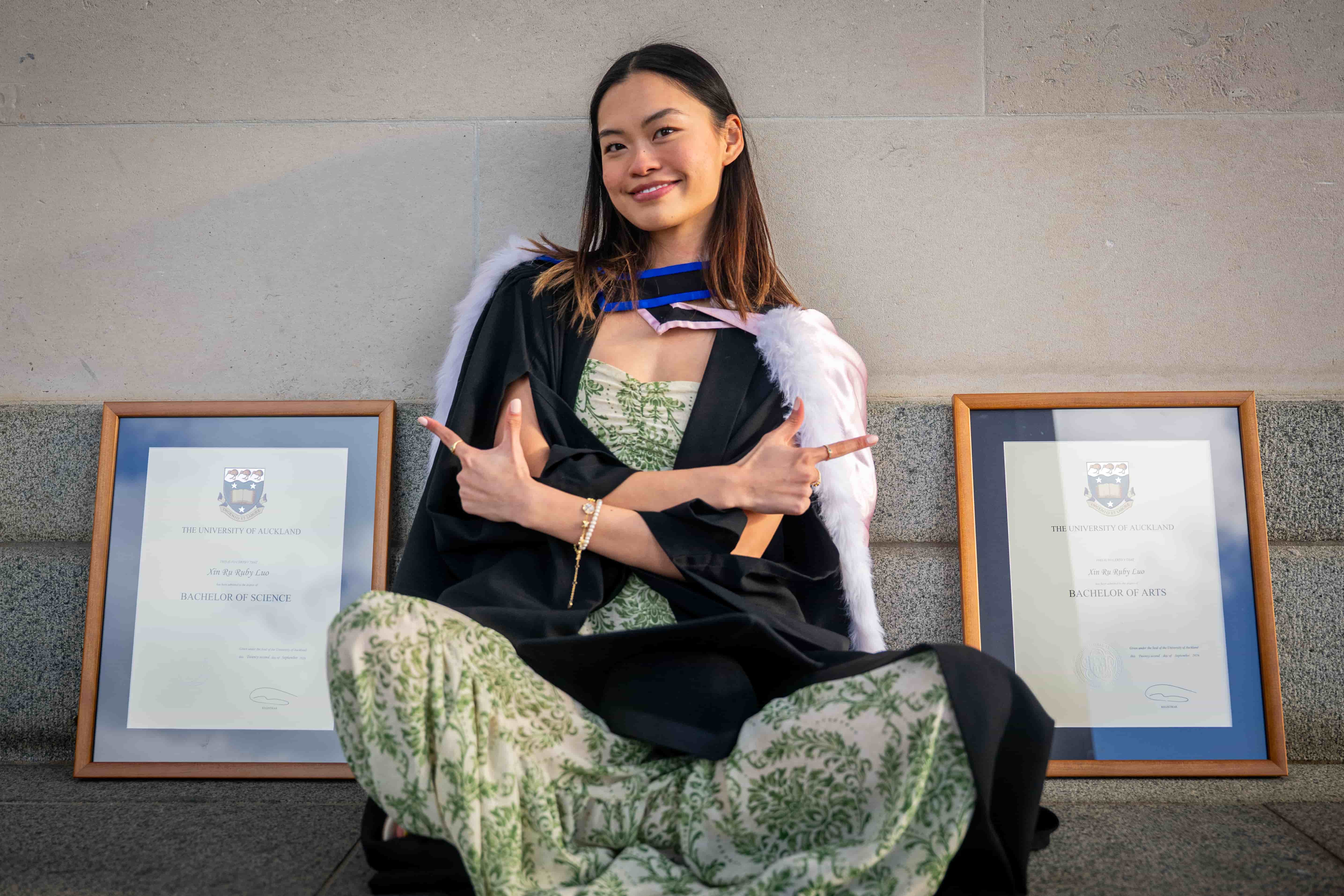 Ruby smiling with arms crossed, sitting between her framed degrees against a stone wall