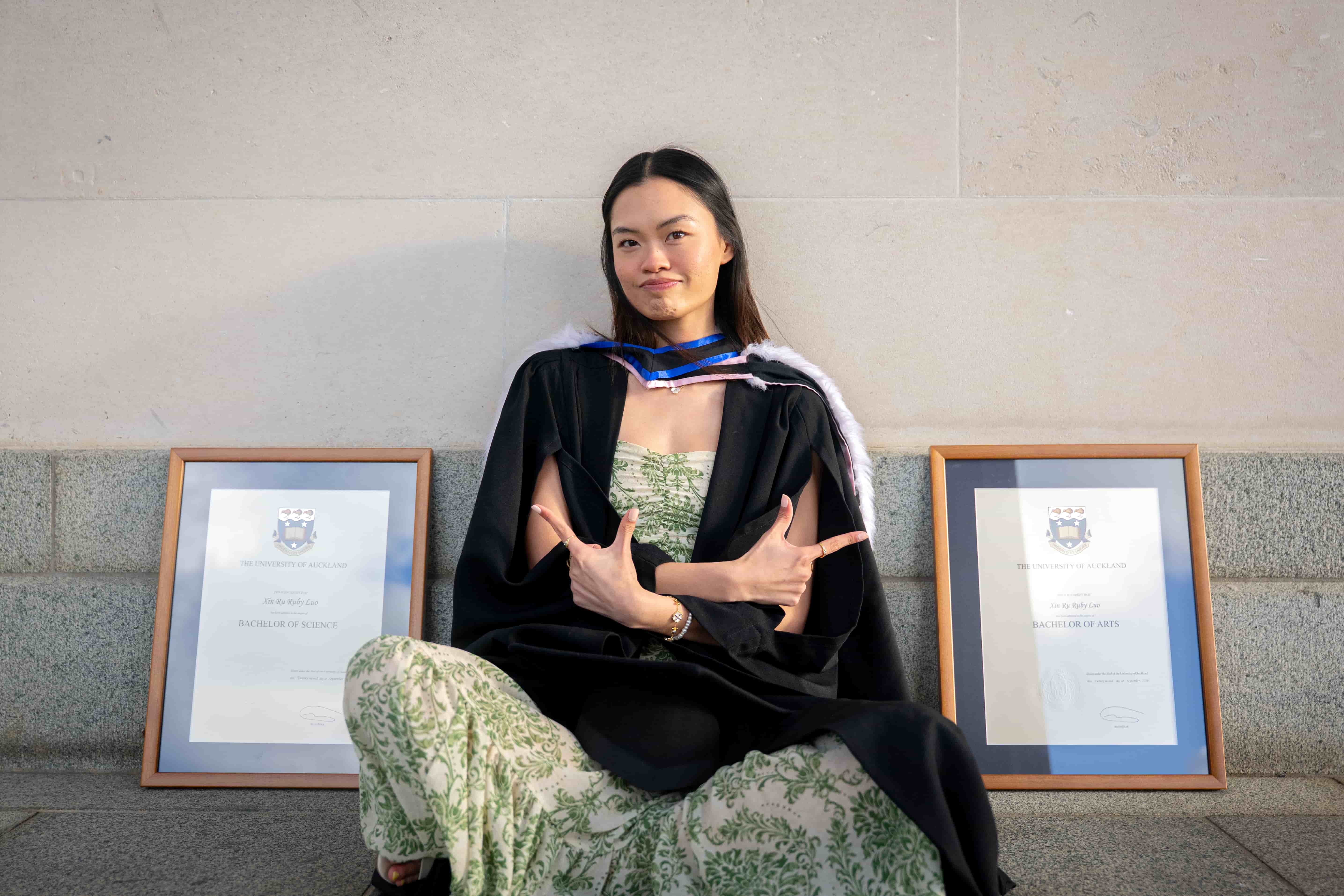 Ruby sitting cross-legged in her graduation gown between her two framed degrees