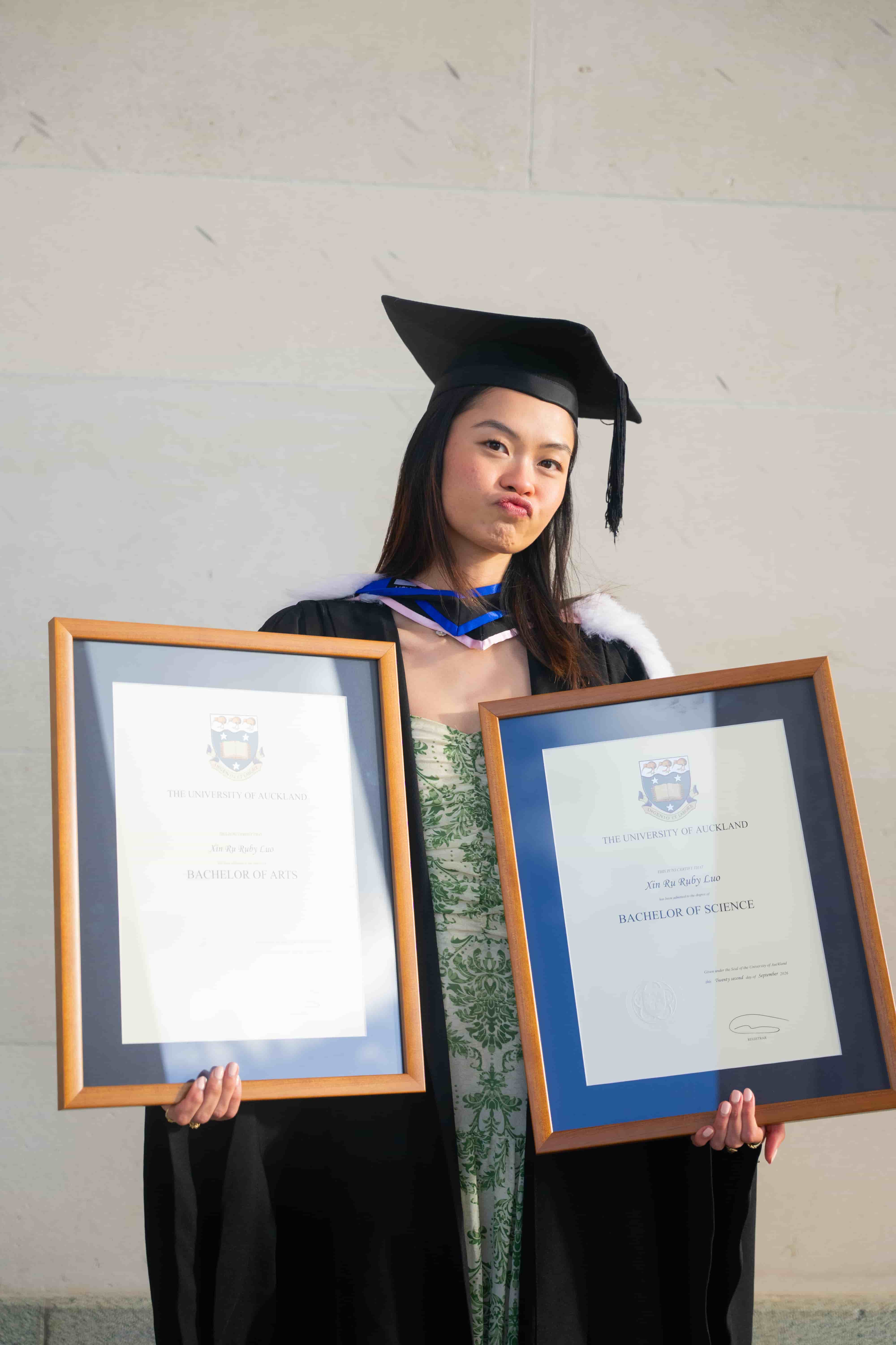 Ruby pulling a playful face while holding both framed degrees against a stone wall