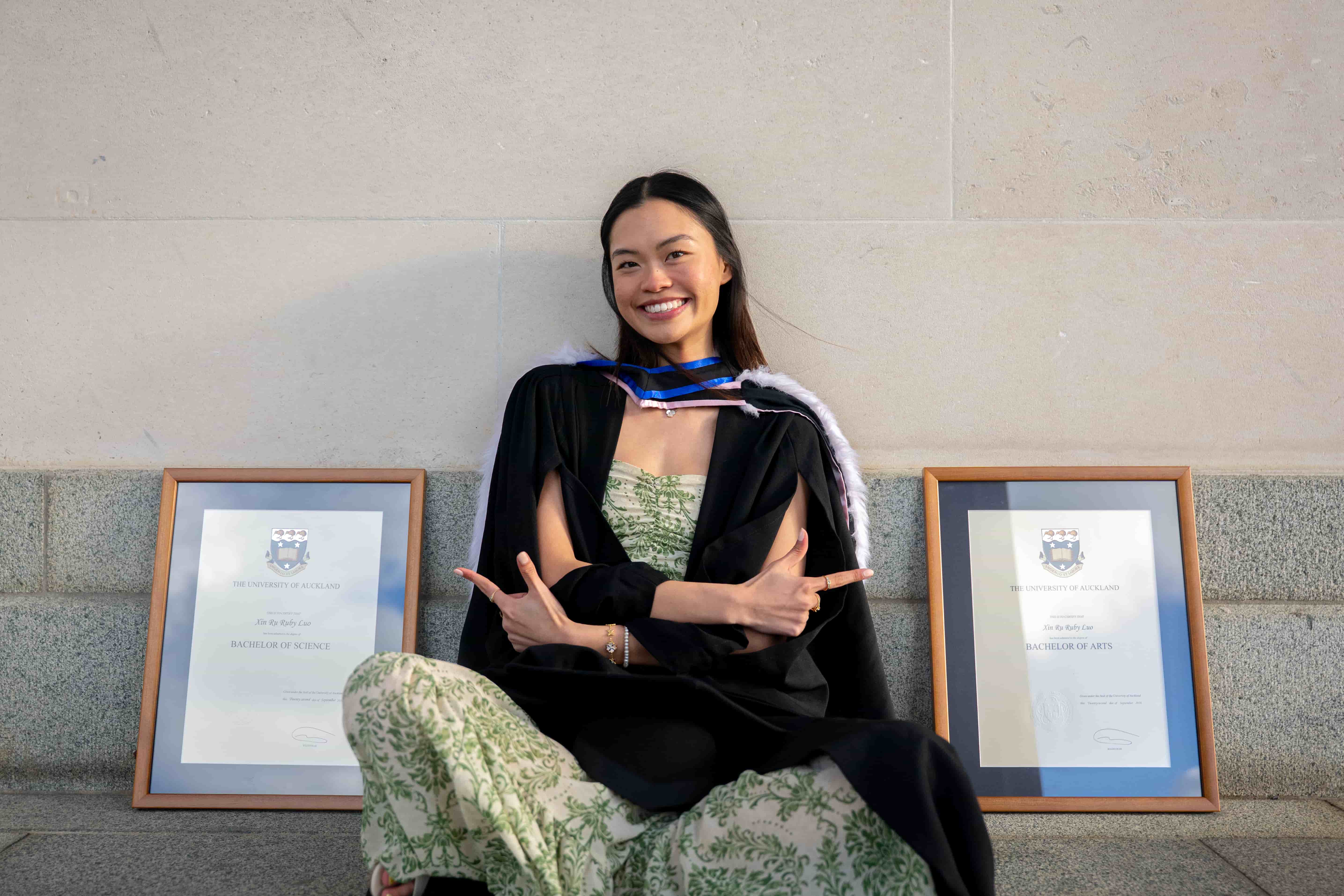 Ruby seated between her framed degrees, arms crossed in a confident pose