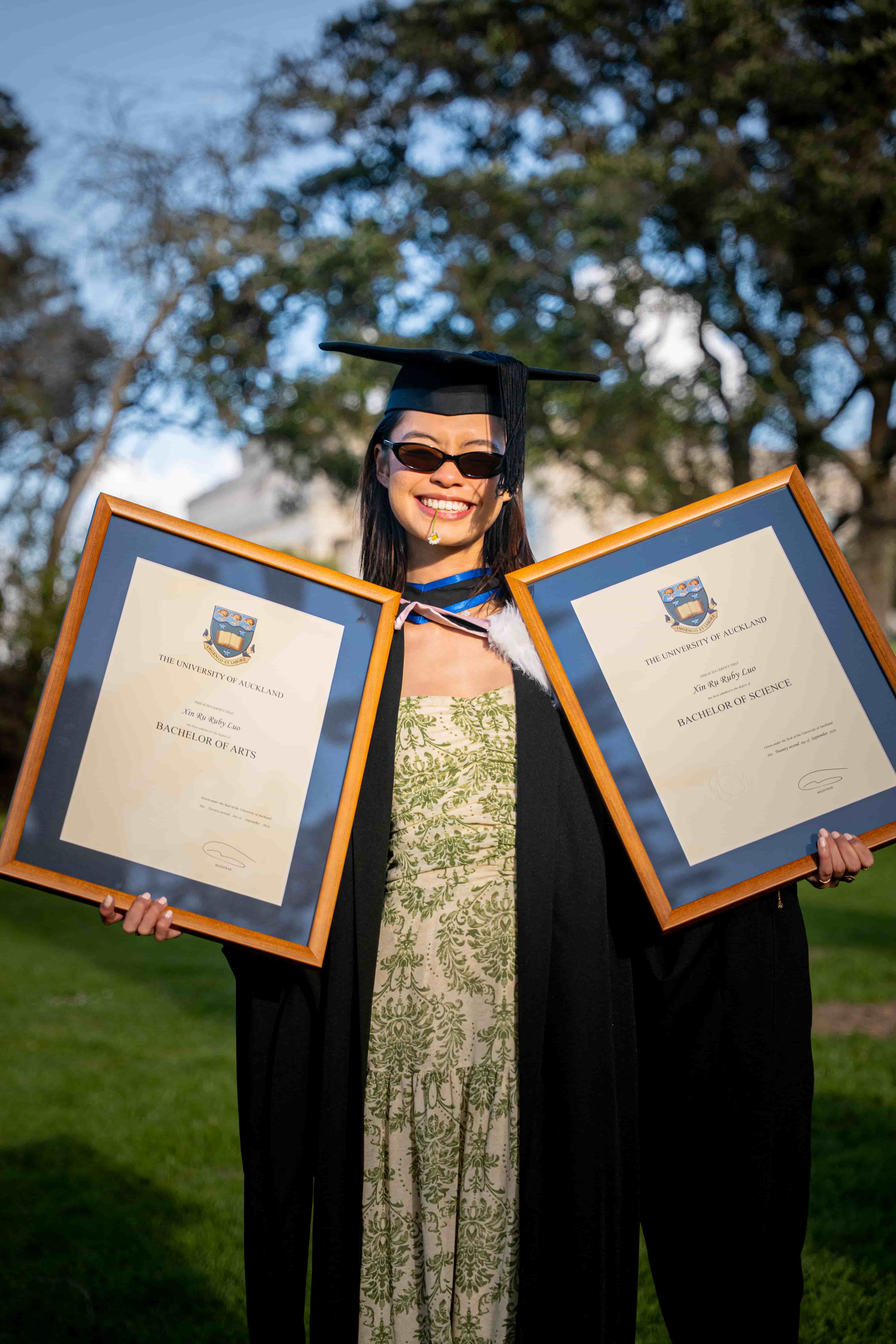 Ruby grinning in sunglasses while holding her two framed degrees on the grass