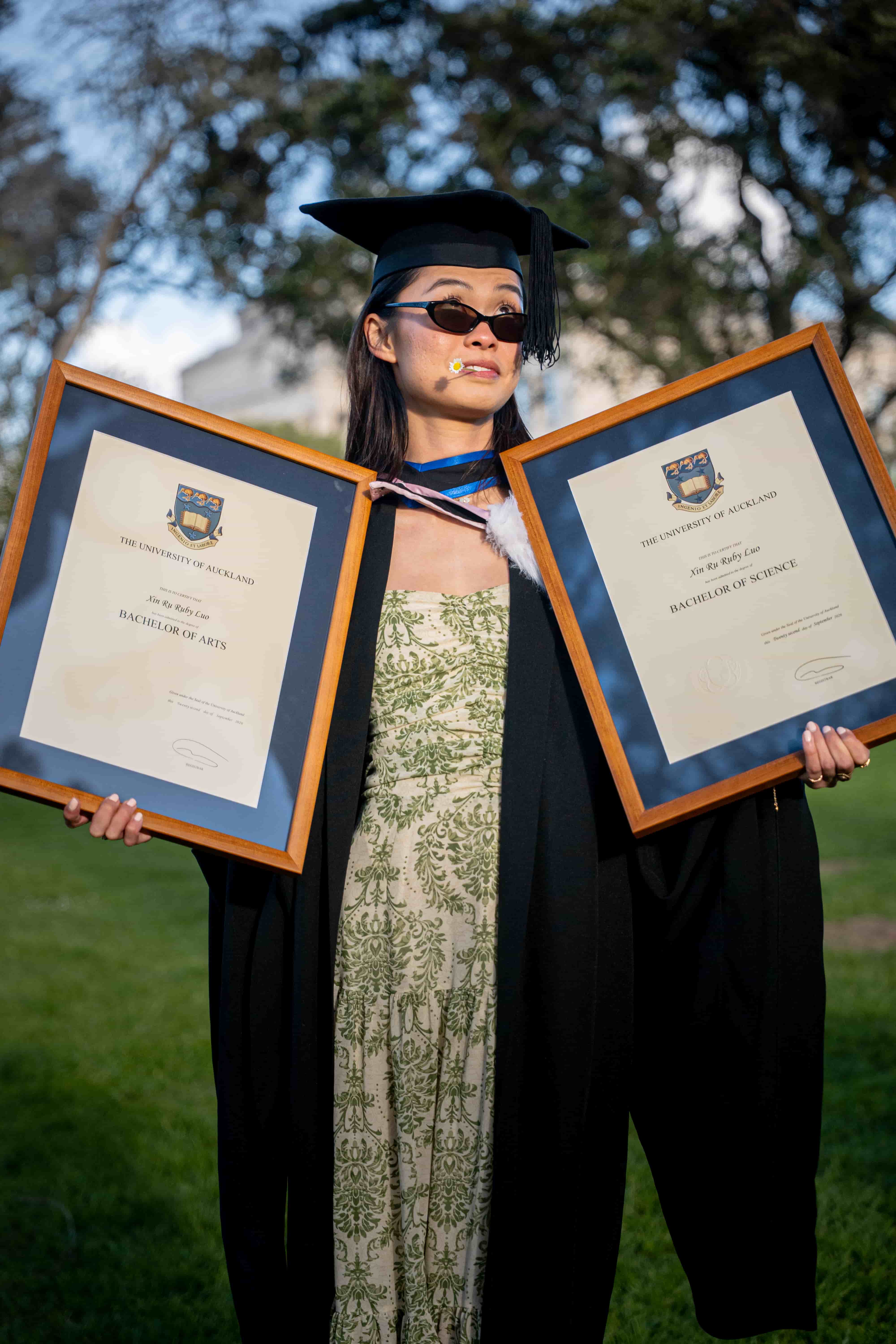 Ruby in sunglasses and graduation cap holding both framed degrees in the Auckland Domain