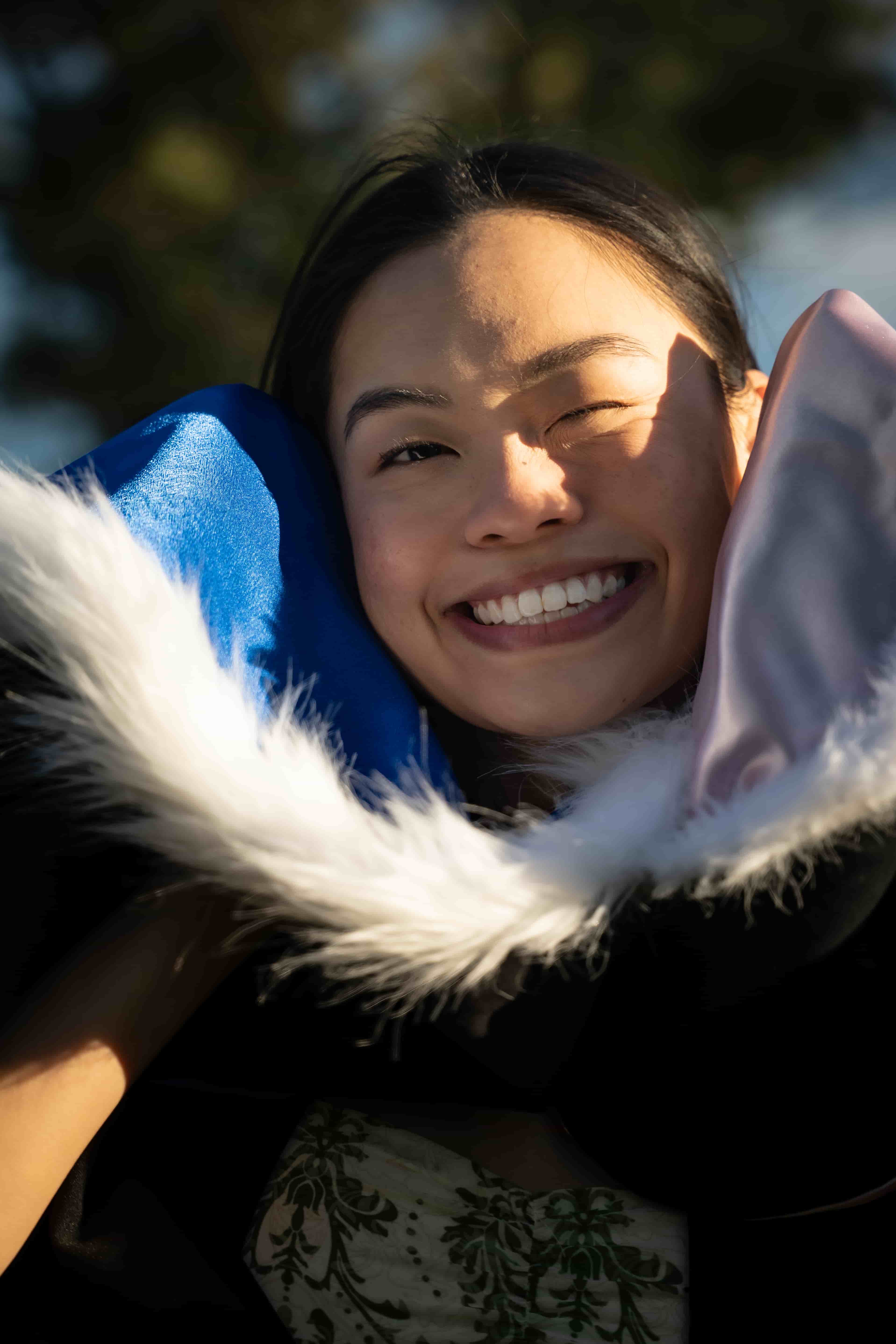 Close-up of Ruby smiling in golden sunlight, wrapped in her blue and pink graduation hood