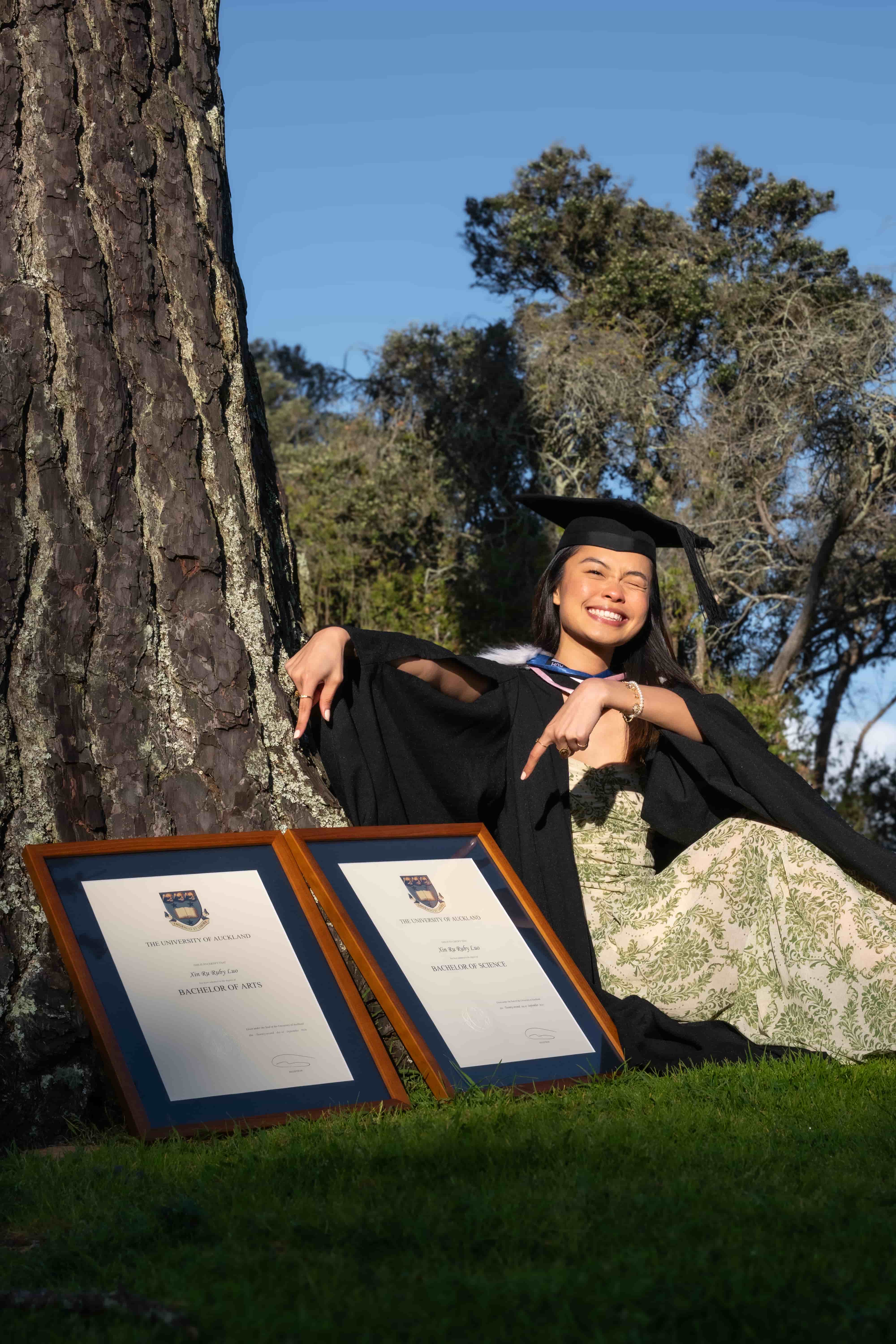 Ruby reclining against a pine tree in her graduation gown beside her framed degrees